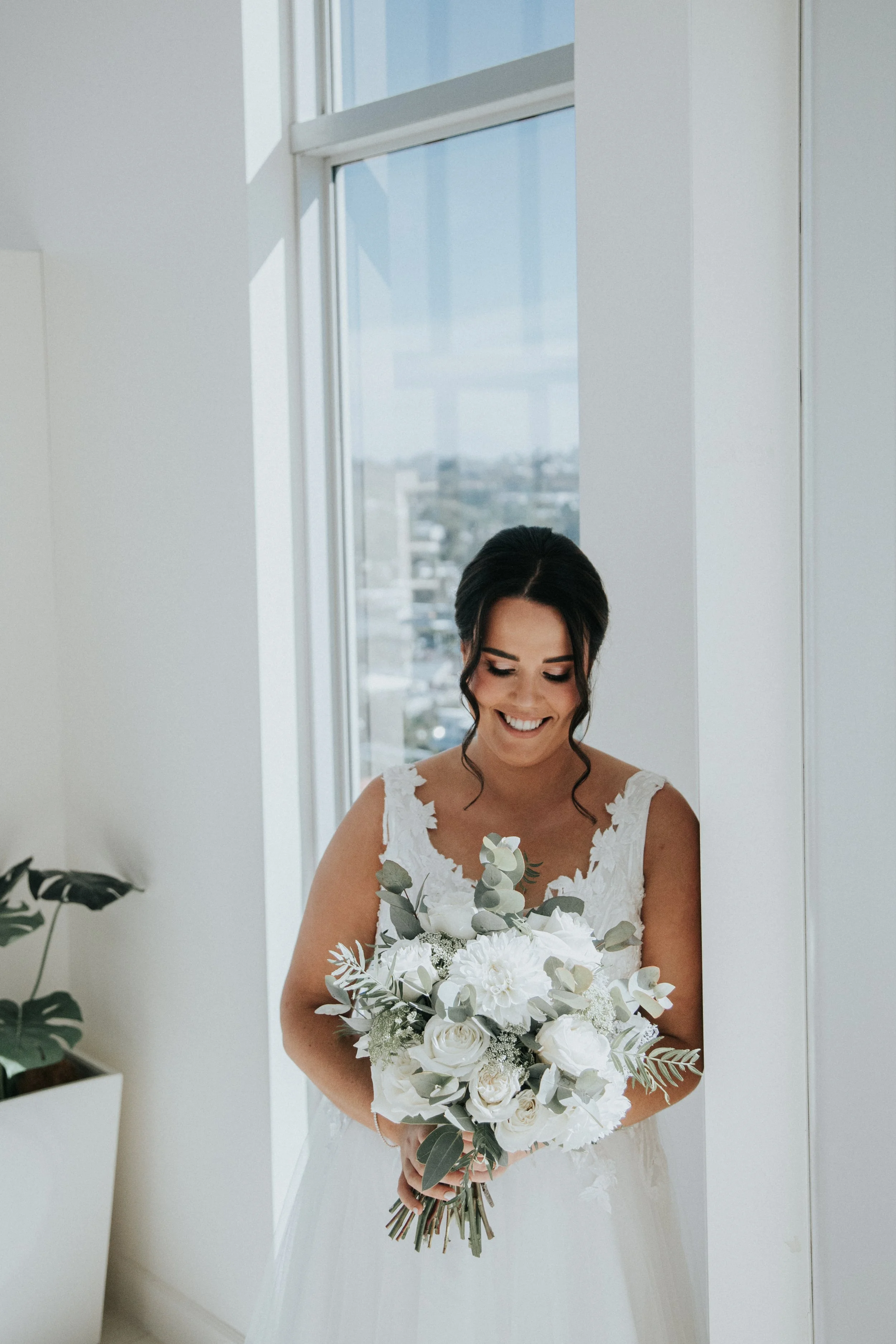 A bride in a white wedding dress holding a bouquet of white flowers, standing by a window with a cityscape view, smiling.