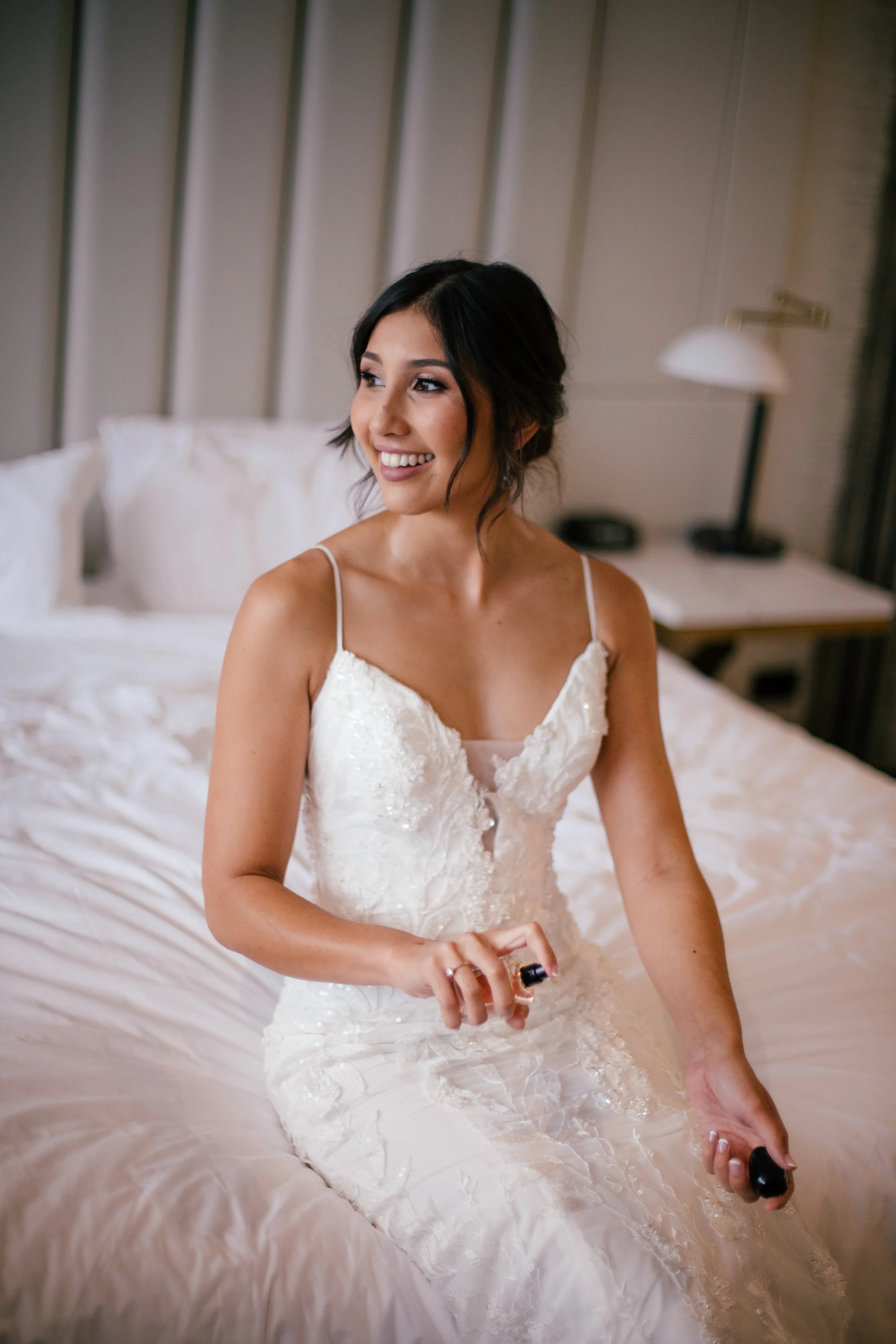A bride in a white wedding dress with thin straps, sitting on a bed and smiling, holding a small object in her right hand.