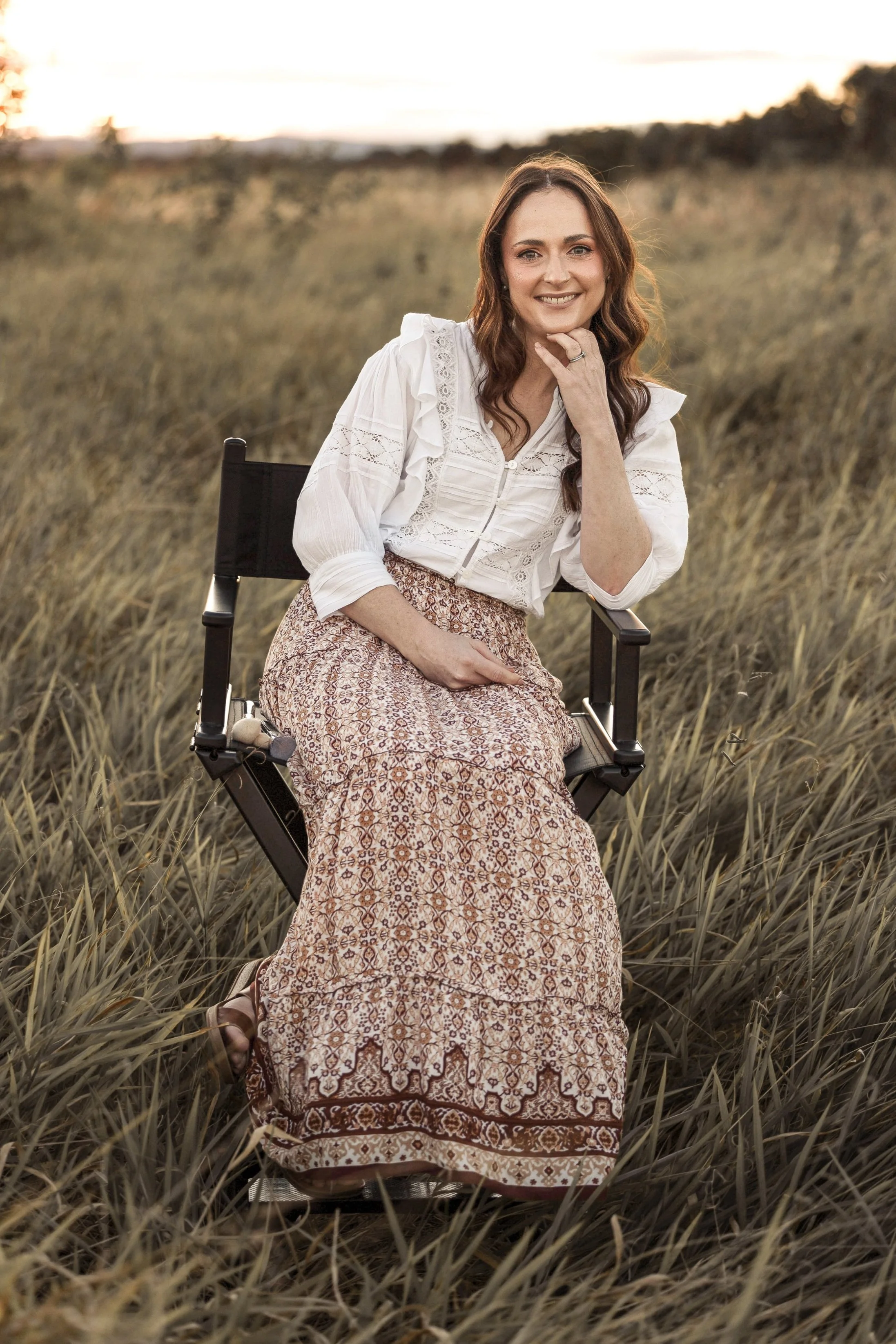A woman sitting on a director's chair in a grassy field during sunset, wearing a white blouse and a patterned maxi skirt, smiling at the camera.
