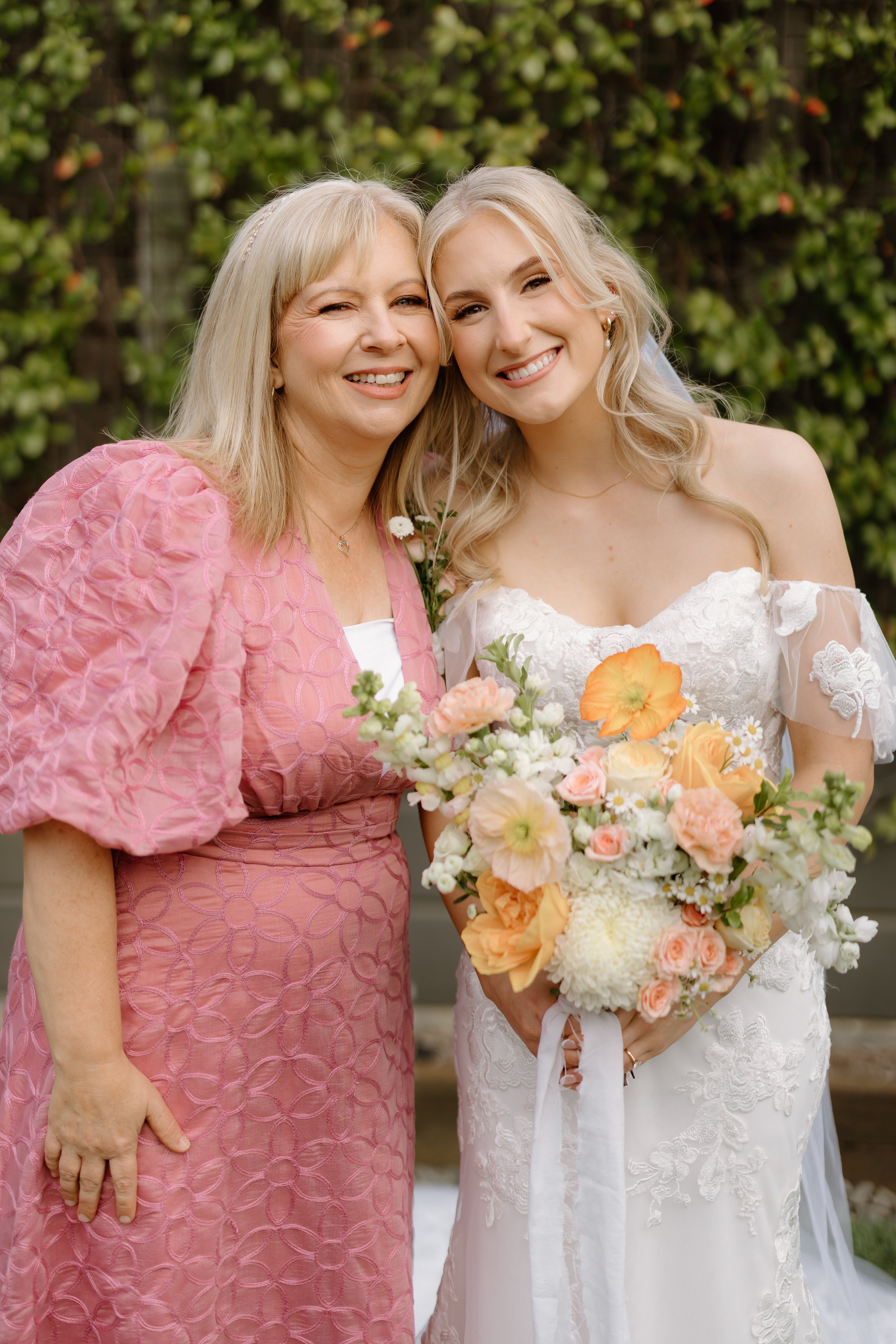 A smiling woman in a pink dress and a smiling woman in a white wedding gown holding a bouquet of pastel-colored flowers, standing outdoors against a background of green foliage.