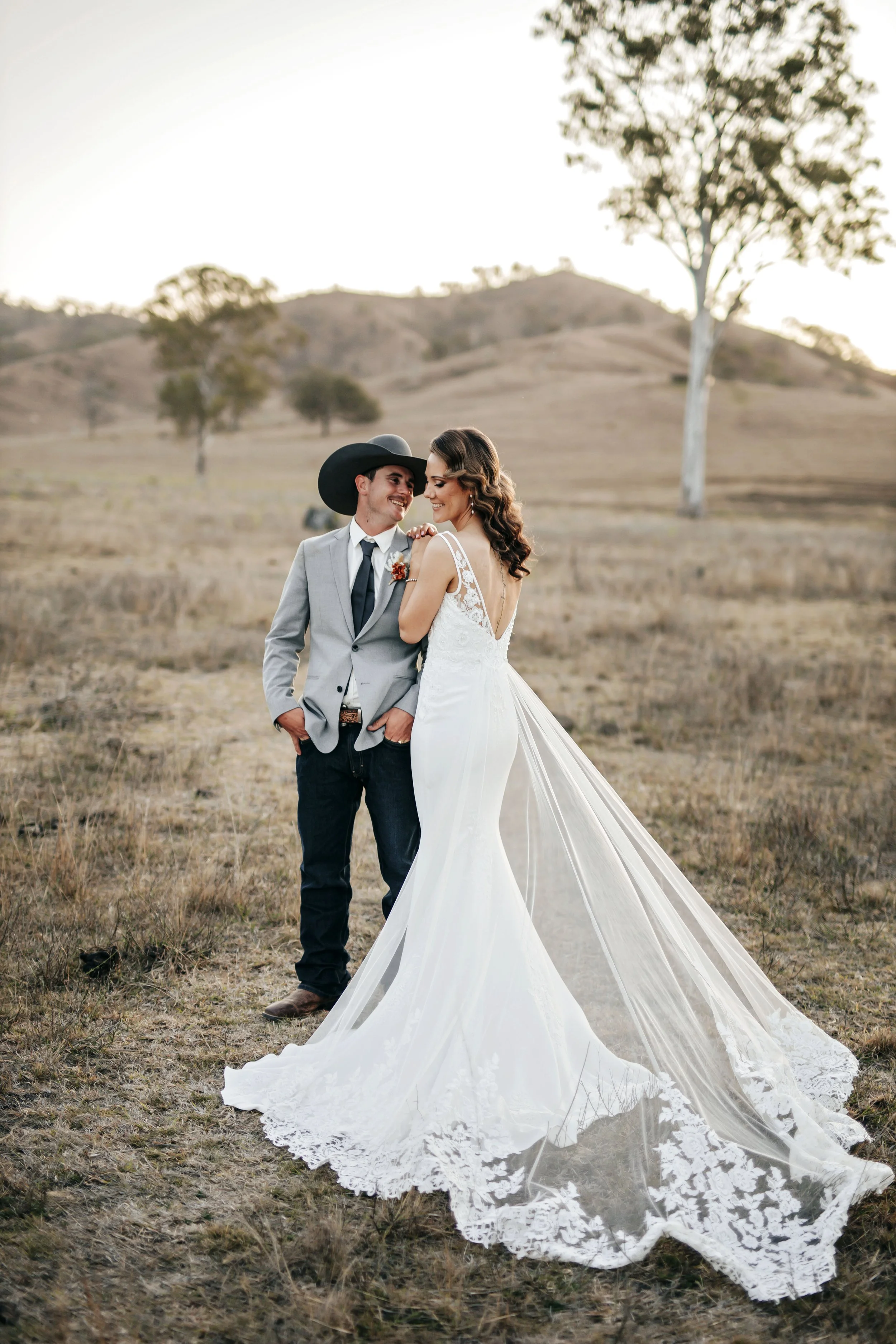 A bride in a white wedding gown and a groom in a gray suit and cowboy hat smile at each other in an outdoor field during sunset.