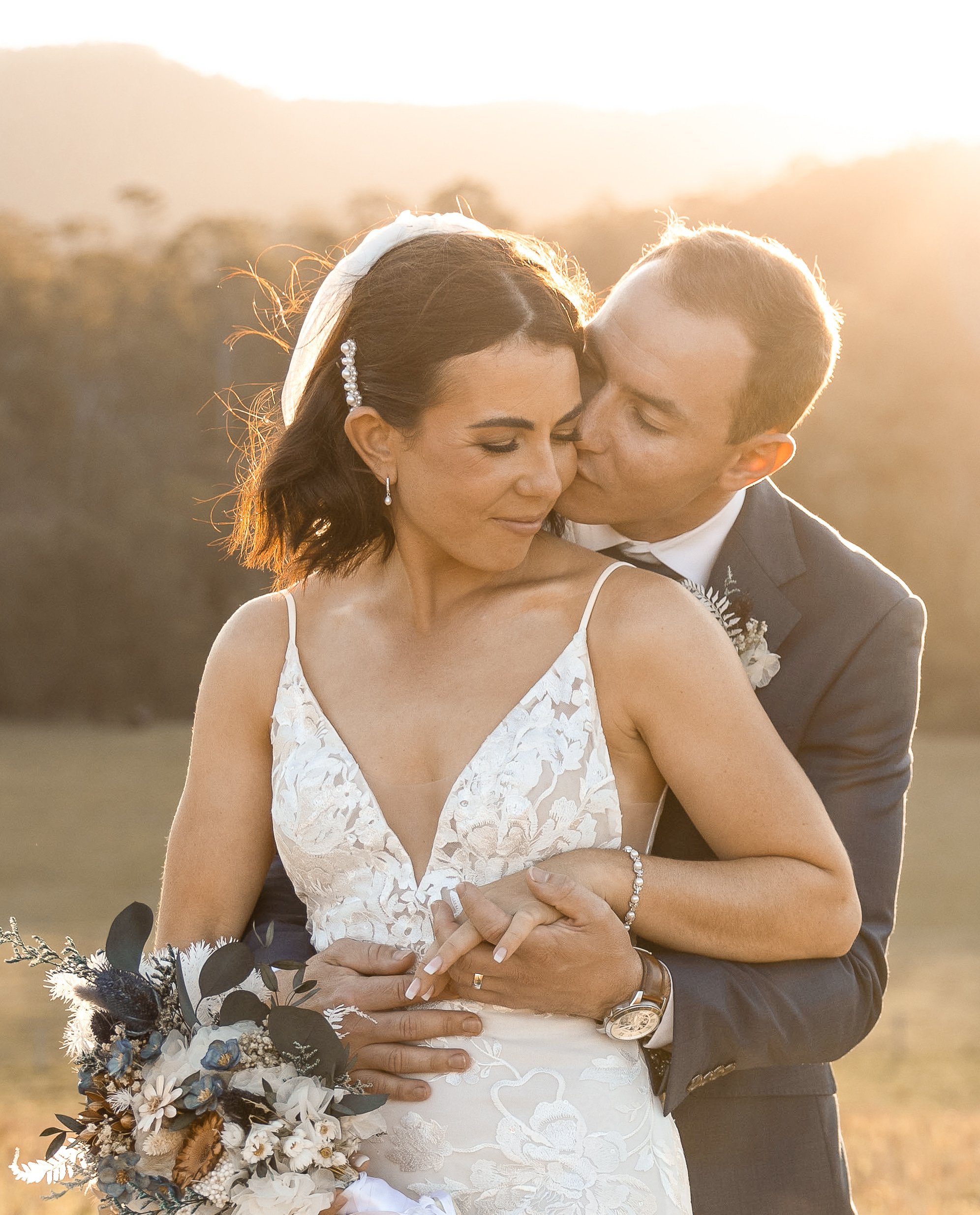 A bride and groom embracing outdoors during sunset, with the groom kissing the bride's cheek.