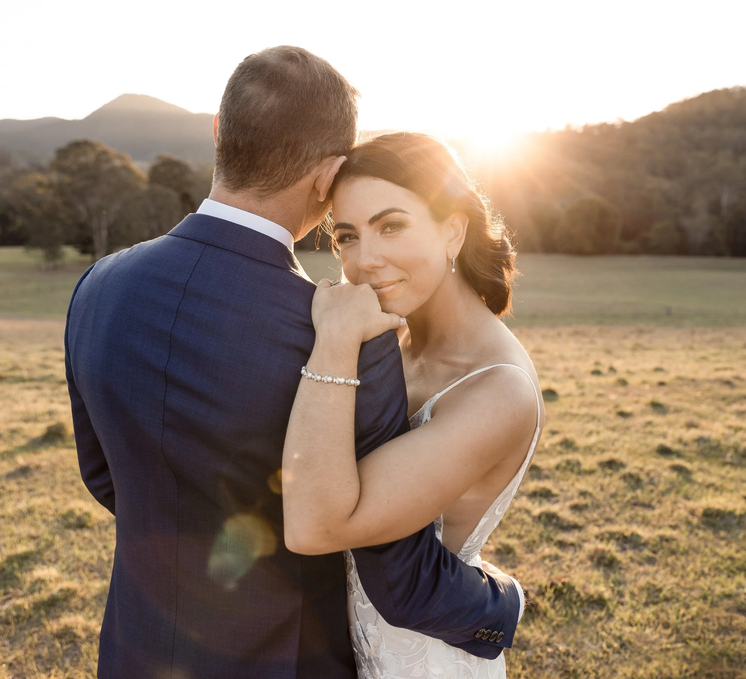 A bride and groom embracing outdoors during sunset with mountains and trees in the background.