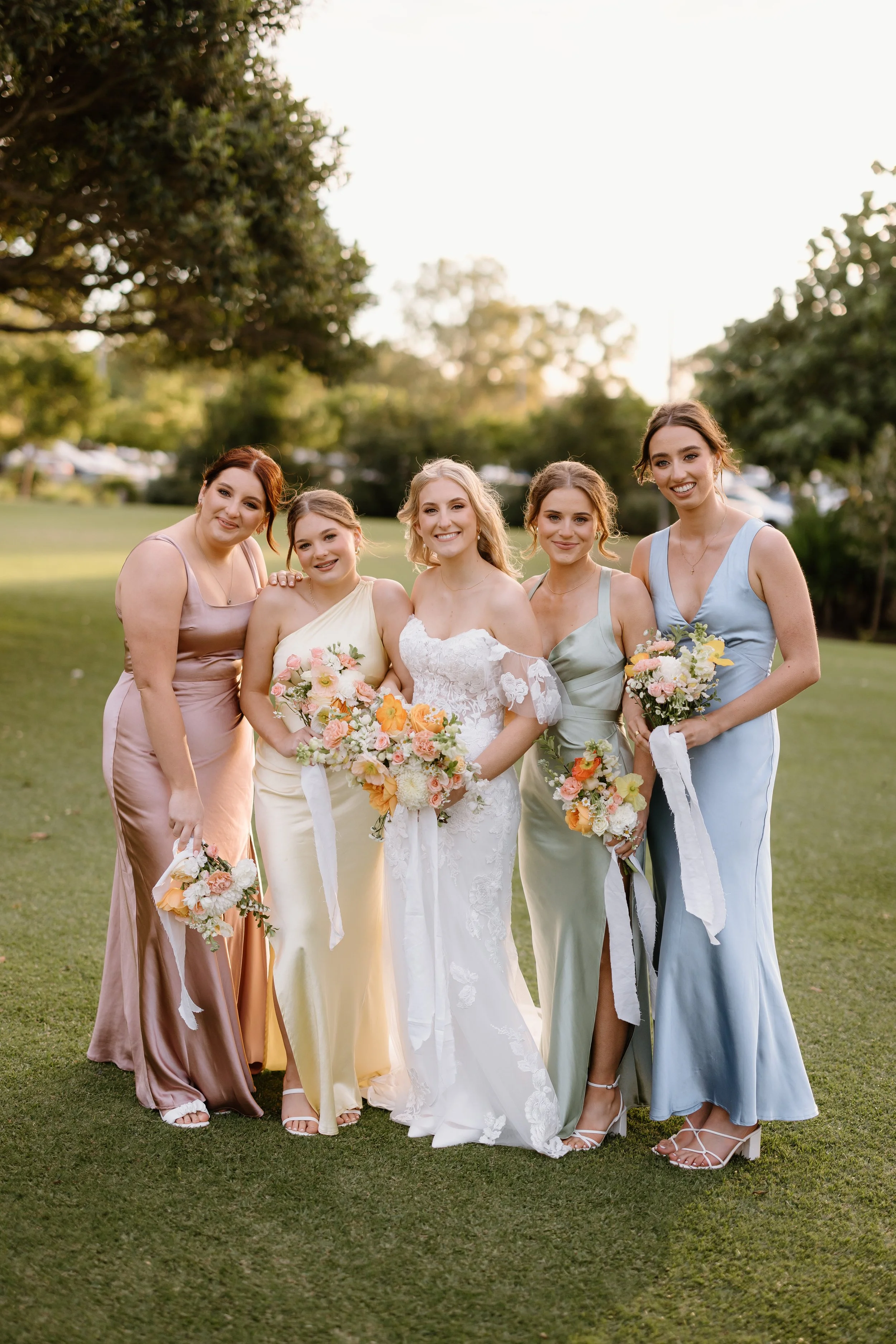A group of five women, including a bride in a white wedding gown, standing outdoors on green grass with trees in the background during sunset. They are smiling and holding bouquets of flowers.