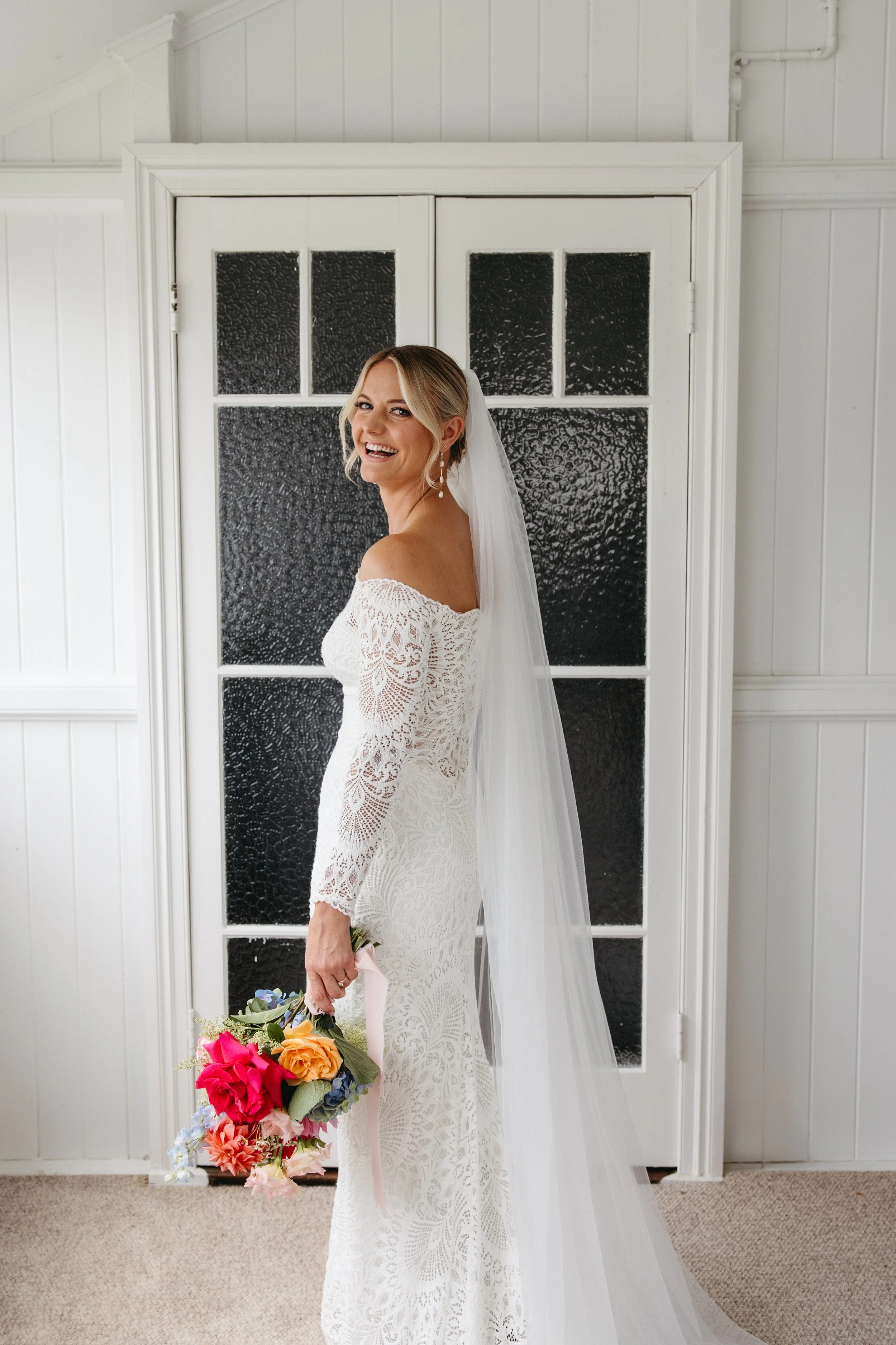 A bride in an off-shoulder lace wedding dress holding a colorful bouquet, smiling and looking back, standing in front of a textured black window with white trim.