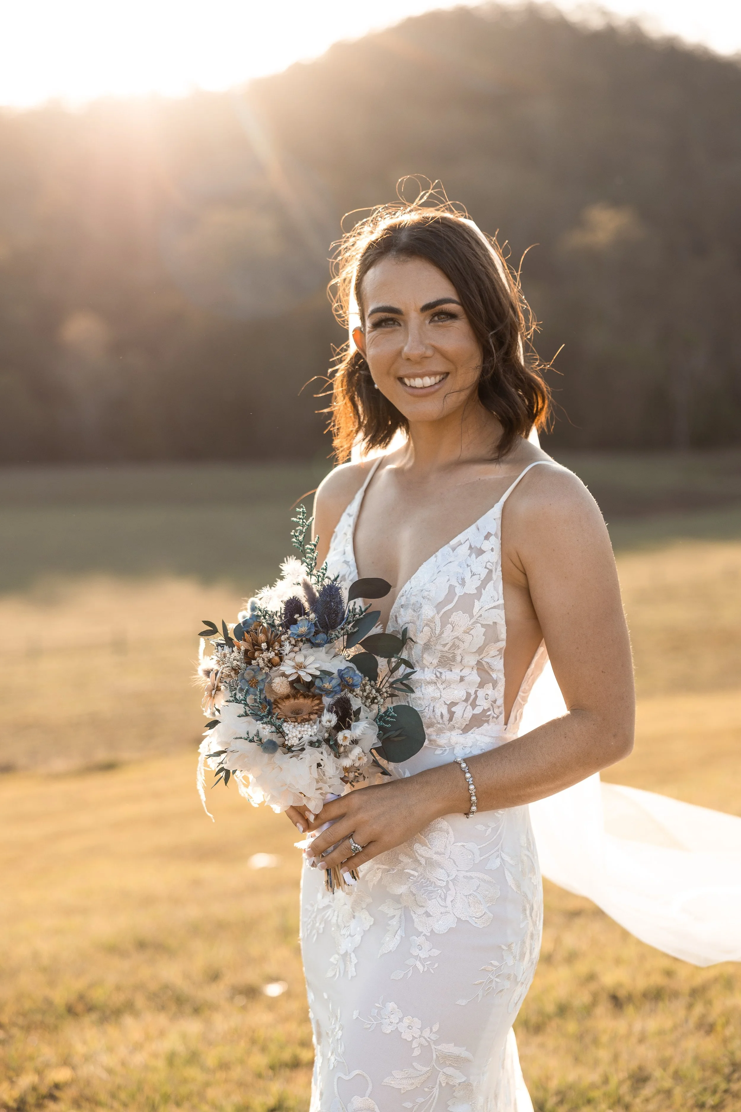 A woman in a white lace wedding dress holding a bouquet of flowers outdoors during sunset, smiling.