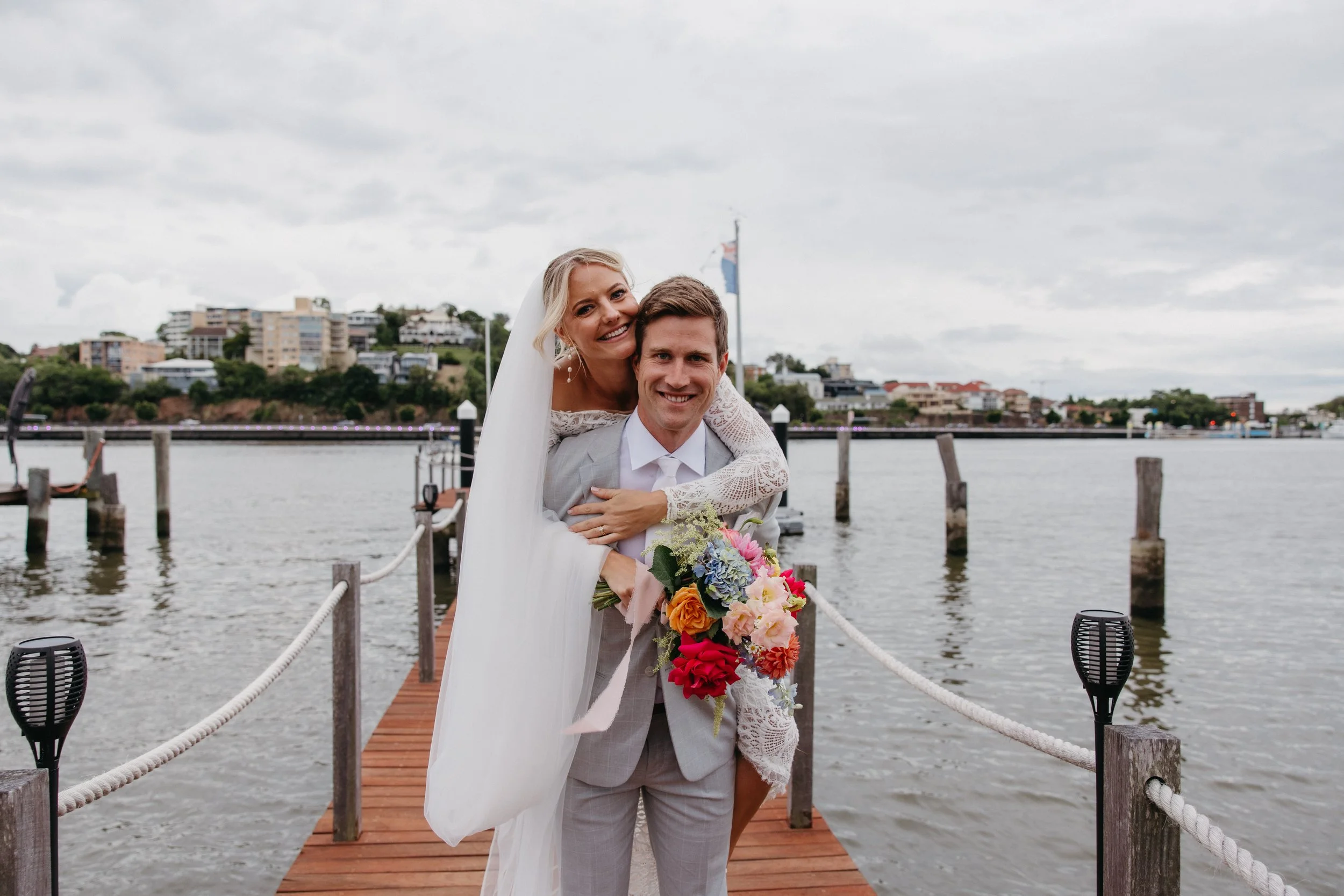 A bride and groom smiling together on a dock by the water, with buildings and cloudy sky in the background. The bride is on the groom's back, holding a colorful bouquet of flowers.