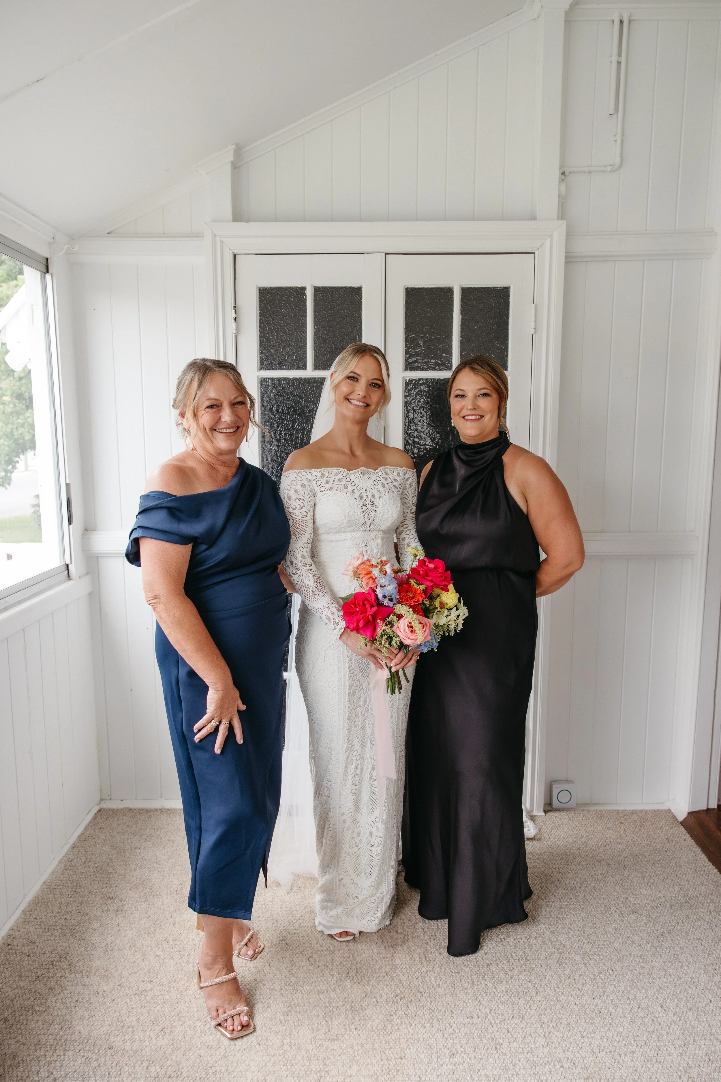 Three women, including a bride in a white lace wedding dress holding a colorful bouquet, standing inside a white room with a door and window.
