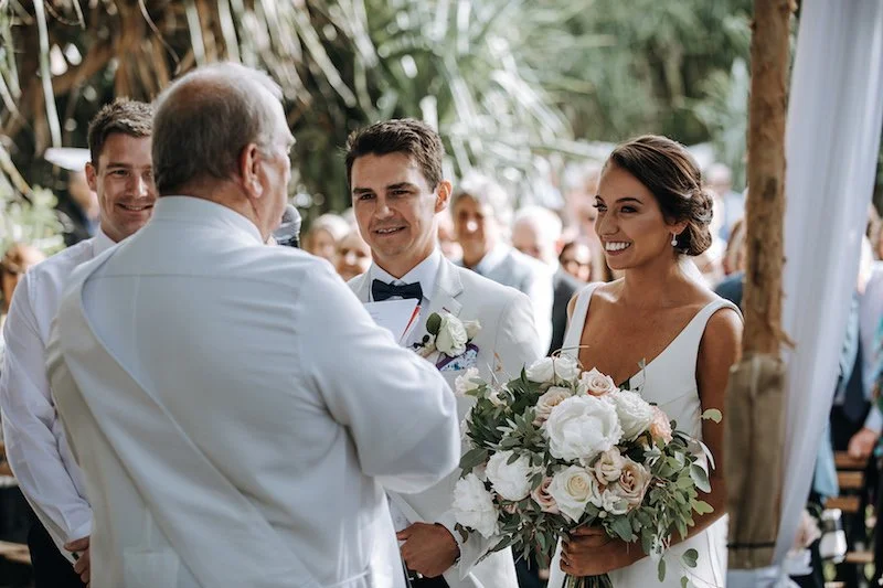 A bride and groom exchanging vows at their wedding ceremony, with the officiant speaking to them, in a tropical outdoor setting surrounded by greenery and guests in the background.