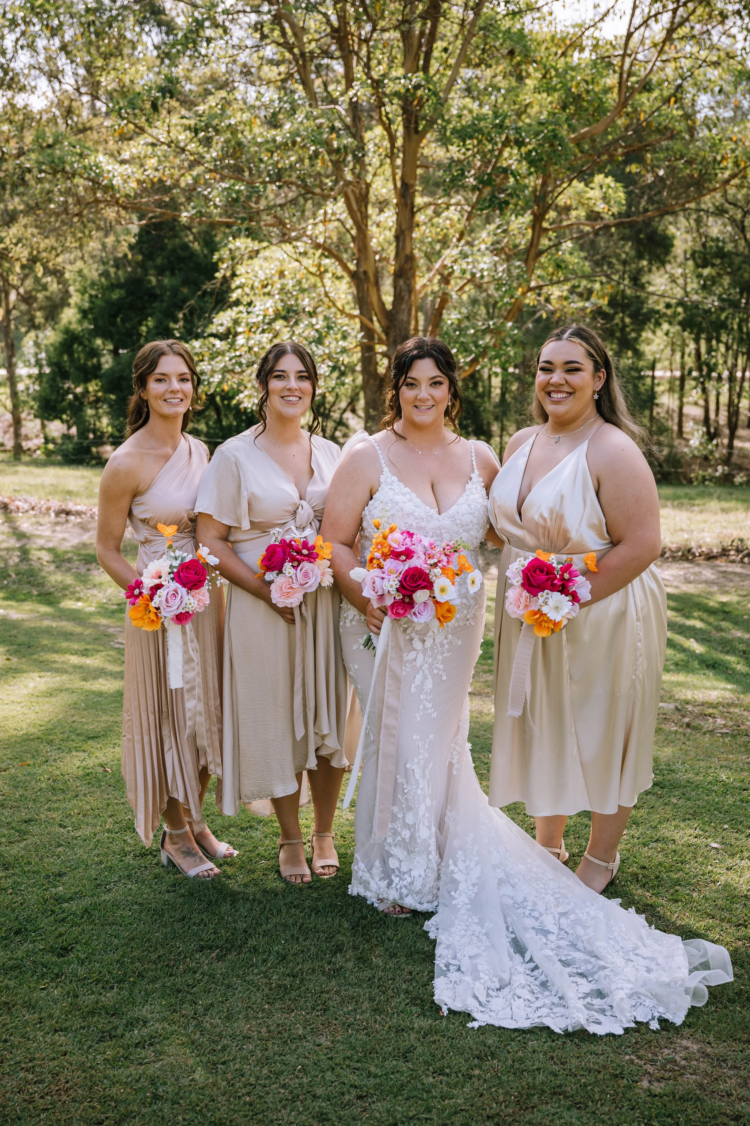 A bride and four bridesmaids standing outdoors on grass, holding colorful bouquets of flowers, with trees in the background on a sunny day.