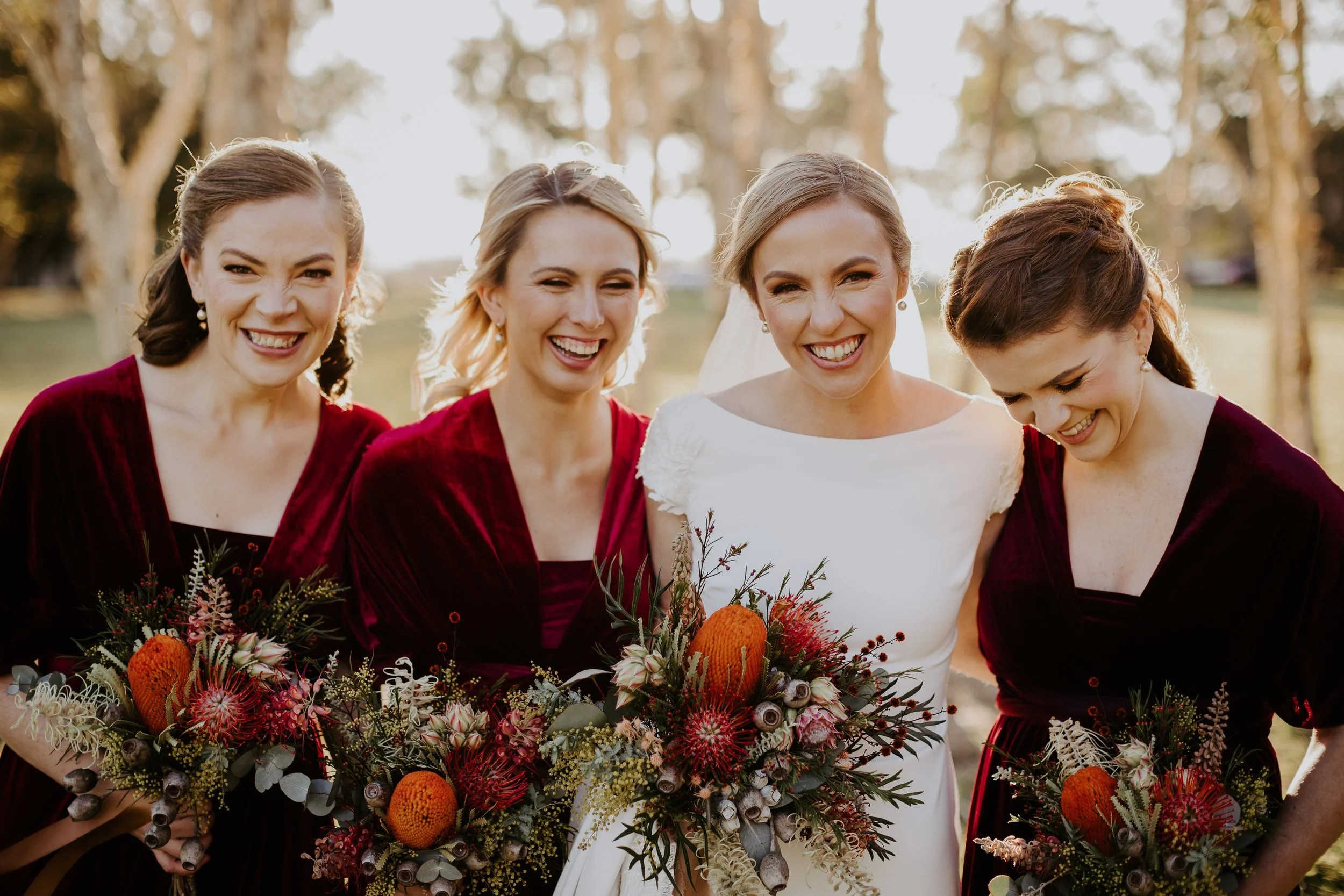Four women, including a bride in a white dress and three bridesmaids in burgundy dresses, smiling and holding bouquets outdoors at sunset.