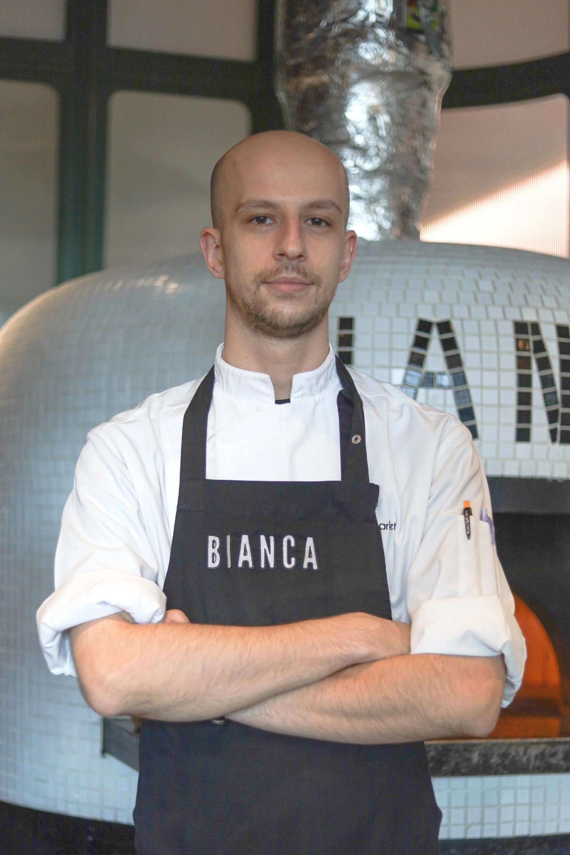 Chef in white uniform and black apron standing with arms crossed in front of a pizza oven.