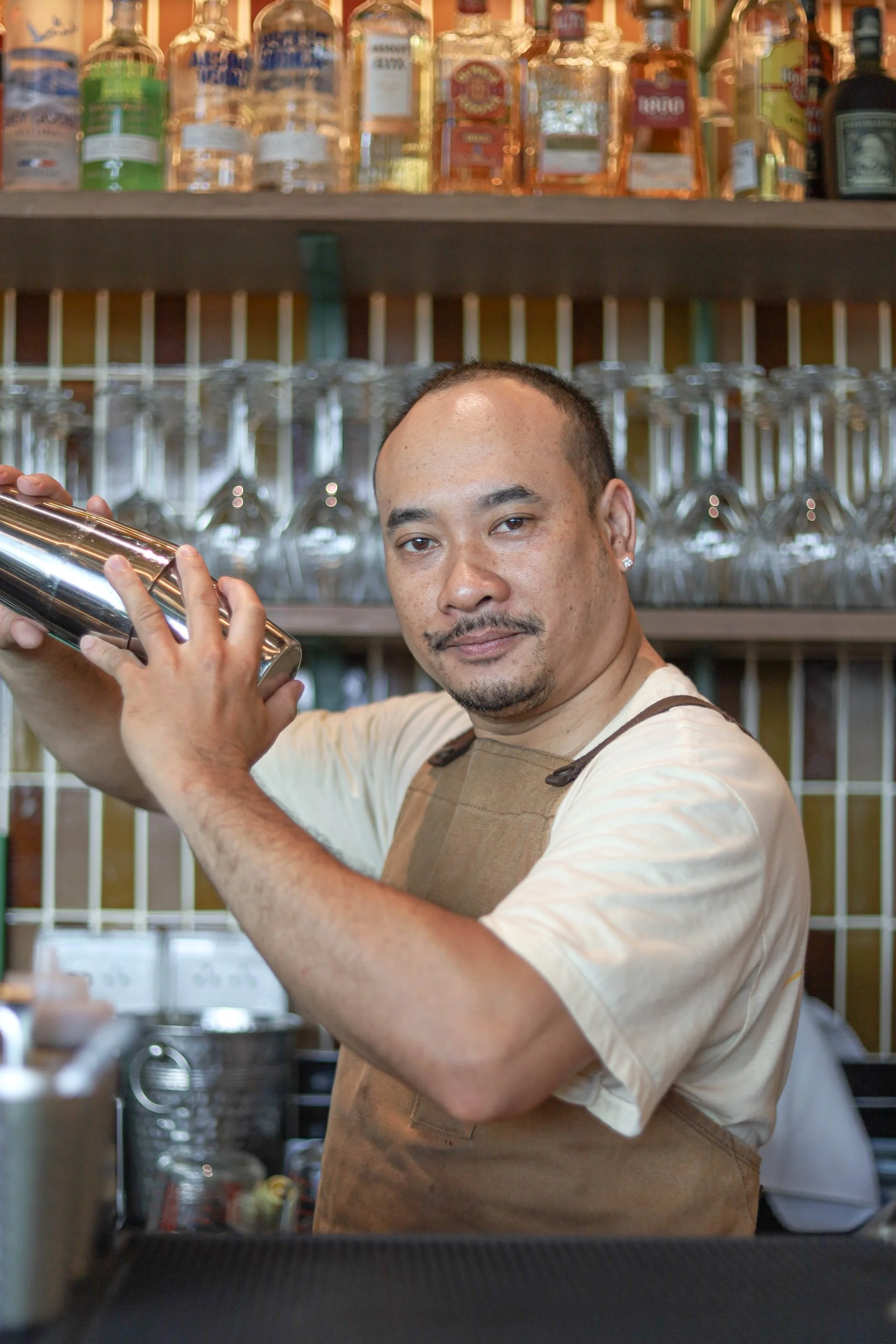 Bartender shaking a cocktail shaker in front of a bar with a shelf of liquor bottles and glassware.