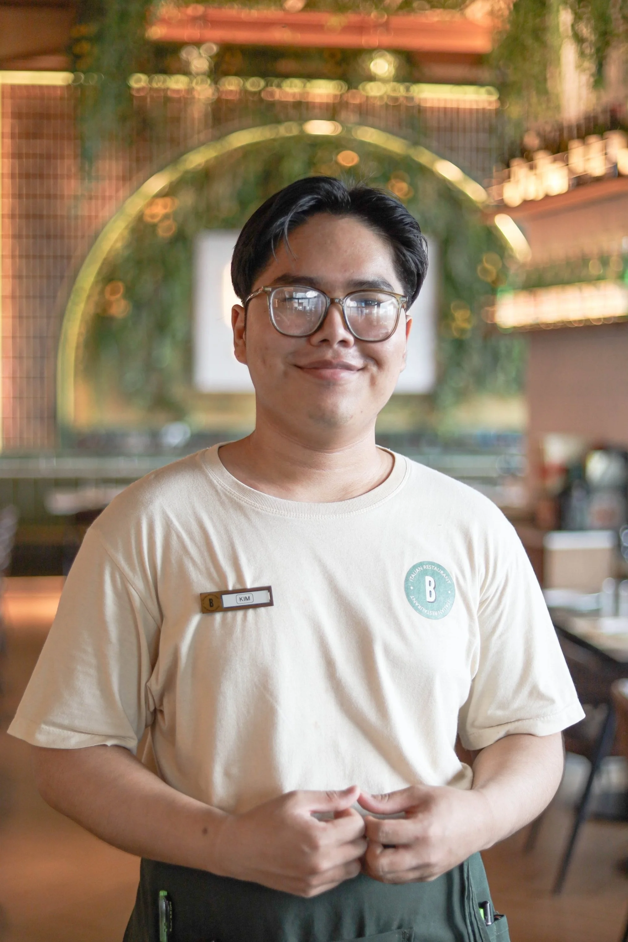 Smiling person in glasses wearing a beige shirt with a name tag standing in a restaurant.