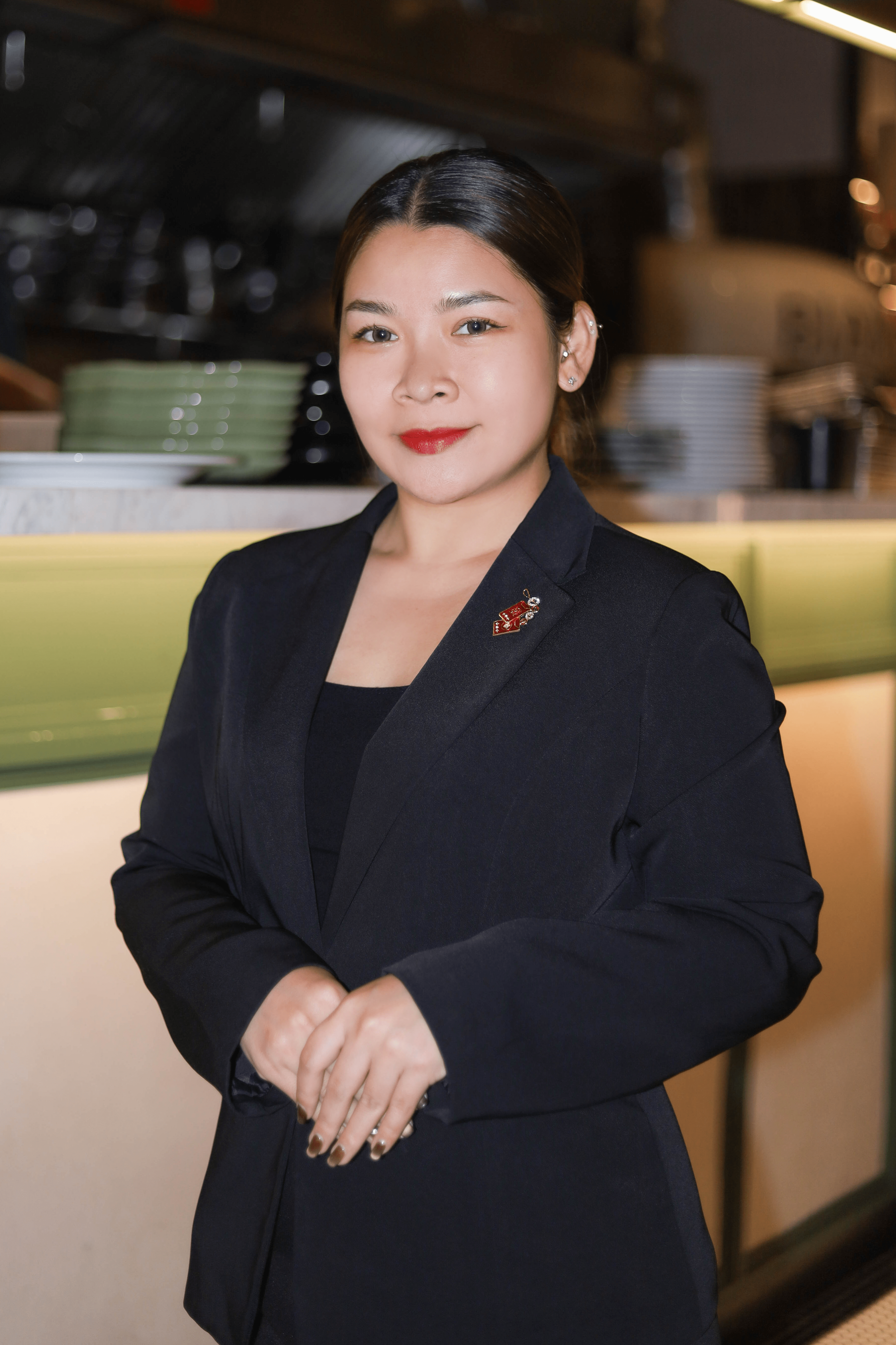 Person in a black suit standing in a restaurant kitchen setting with plates in the background.