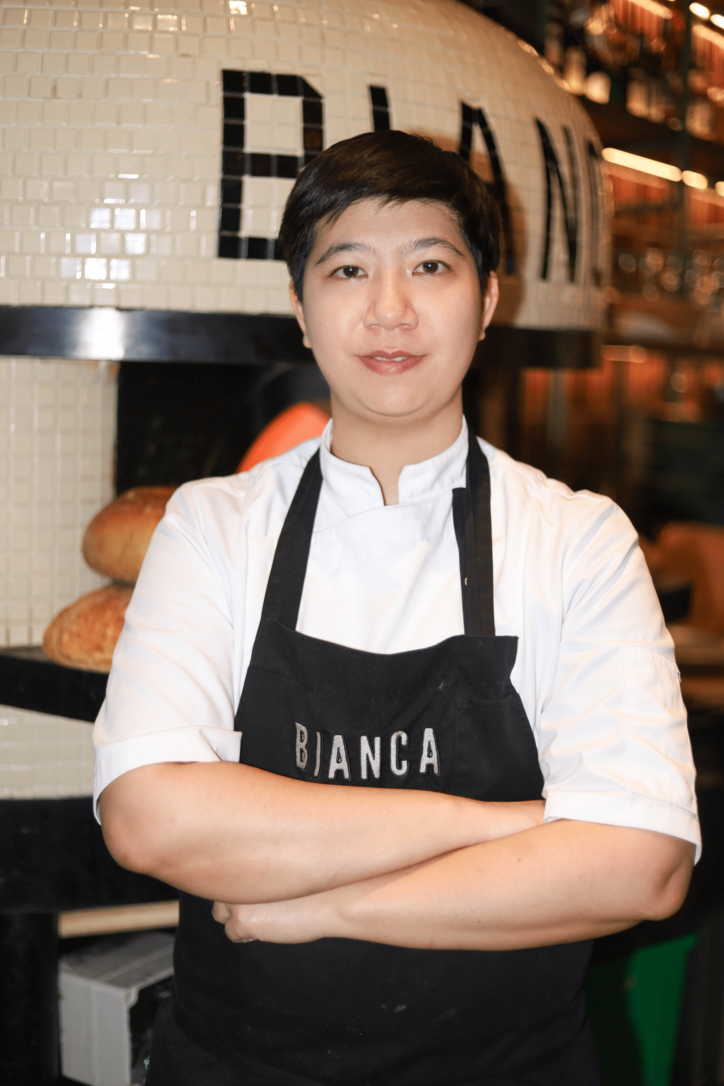 Chef standing in front of a pizza oven with "BIANCA" on apron and oven, arms crossed.