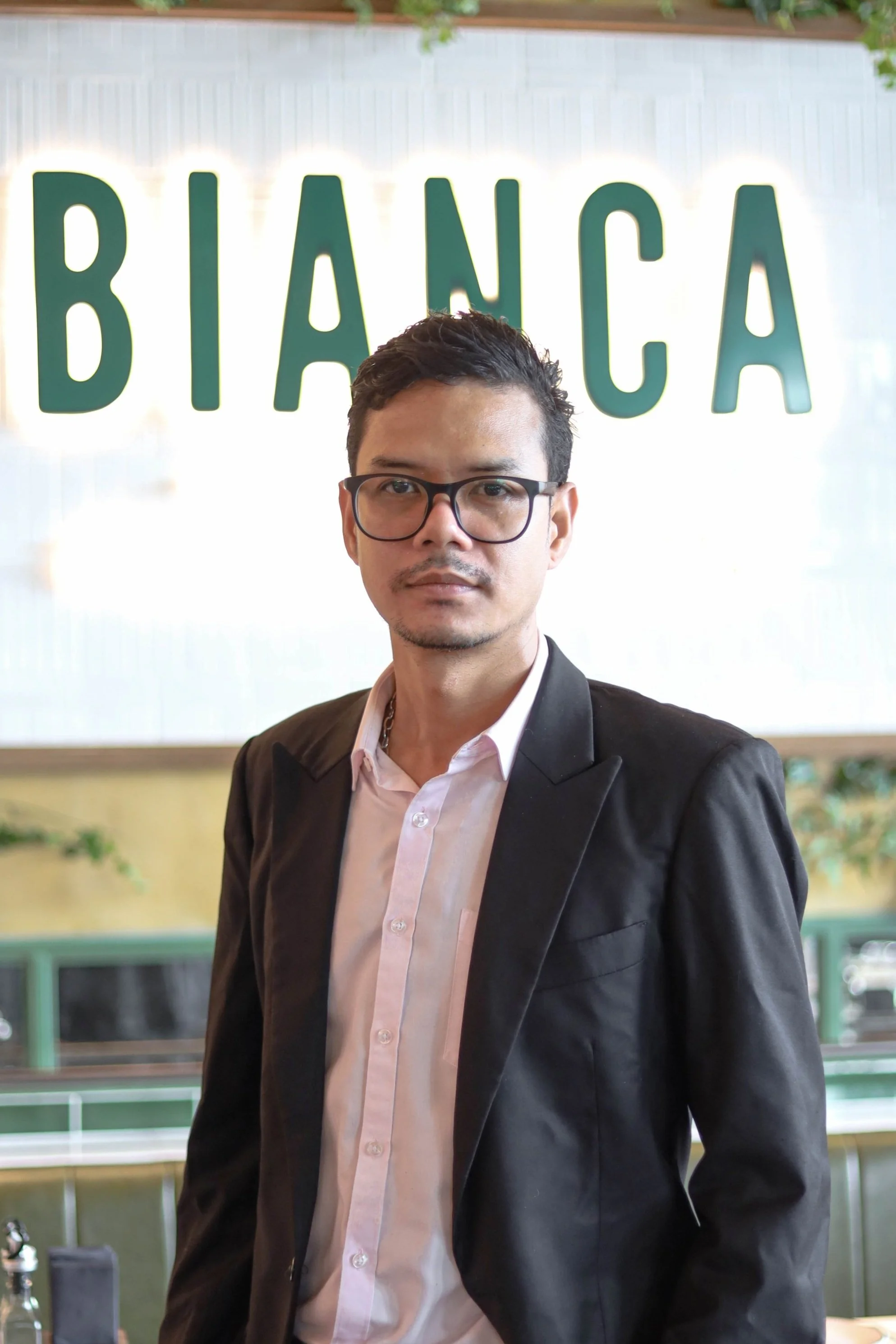 Person wearing glasses and a suit jacket in front of a "BIANCA" sign.