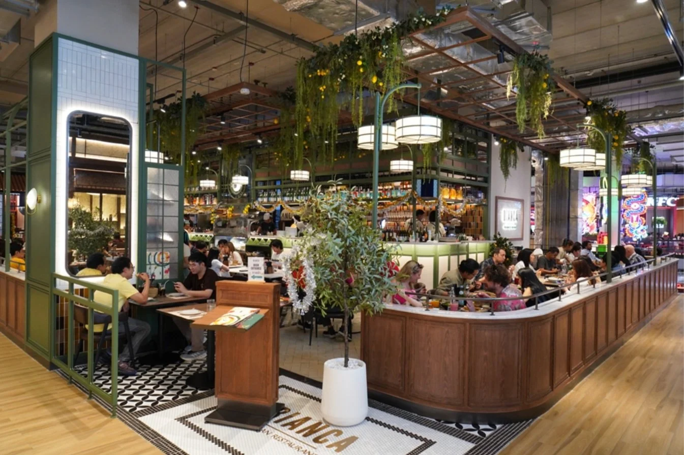 Interior of a modern restaurant with people dining, greenery hanging from the ceiling, and ambient lighting. The restaurant features a mix of wooden and tiled flooring, and a bar area in the background. It has a casual, contemporary design with plants accentuating the decor.