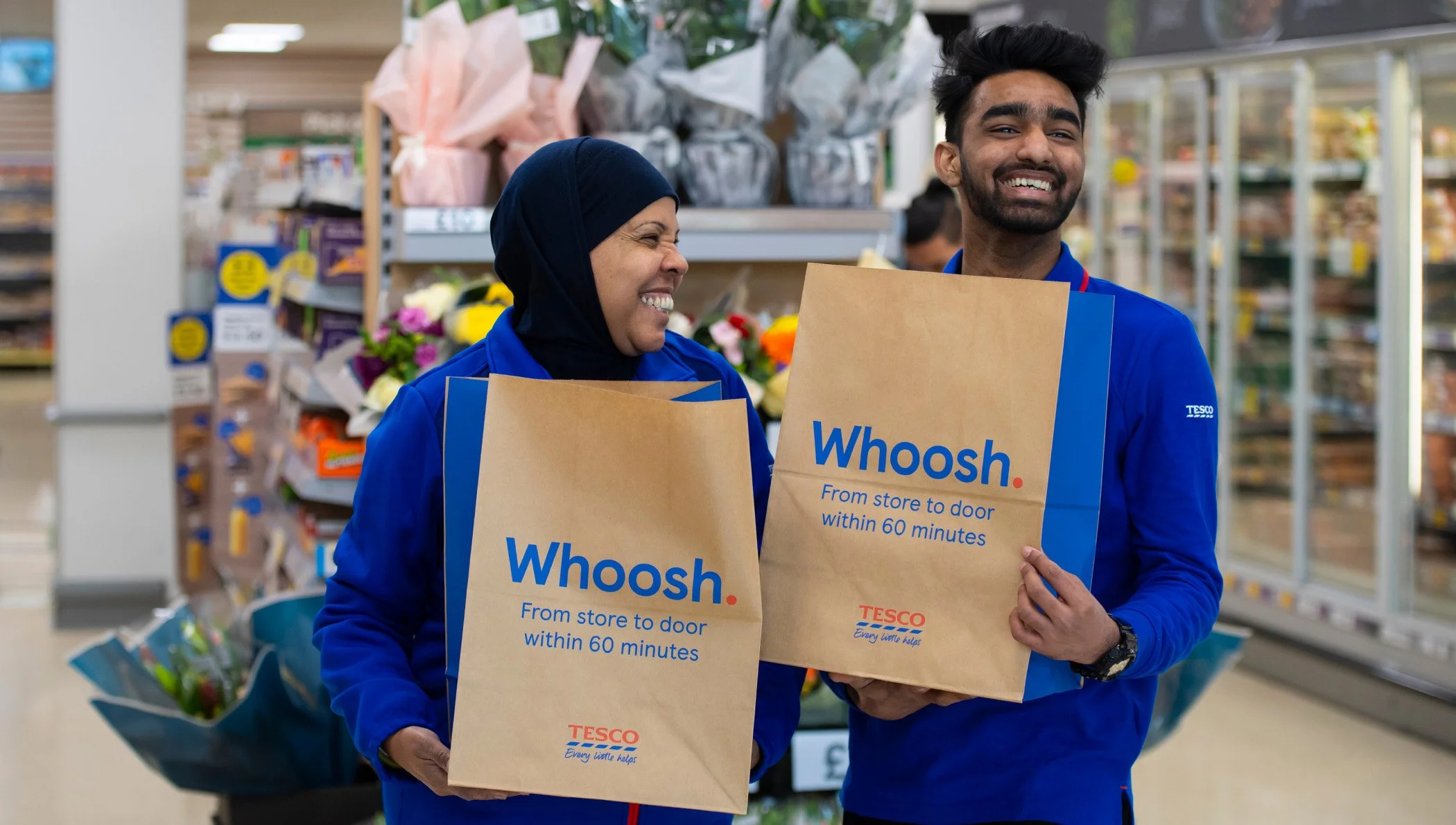 Two Tesco employees smiling and holding shopping bags that read 'Whoosh. From store to door within 60 minutes,' inside a grocery store.