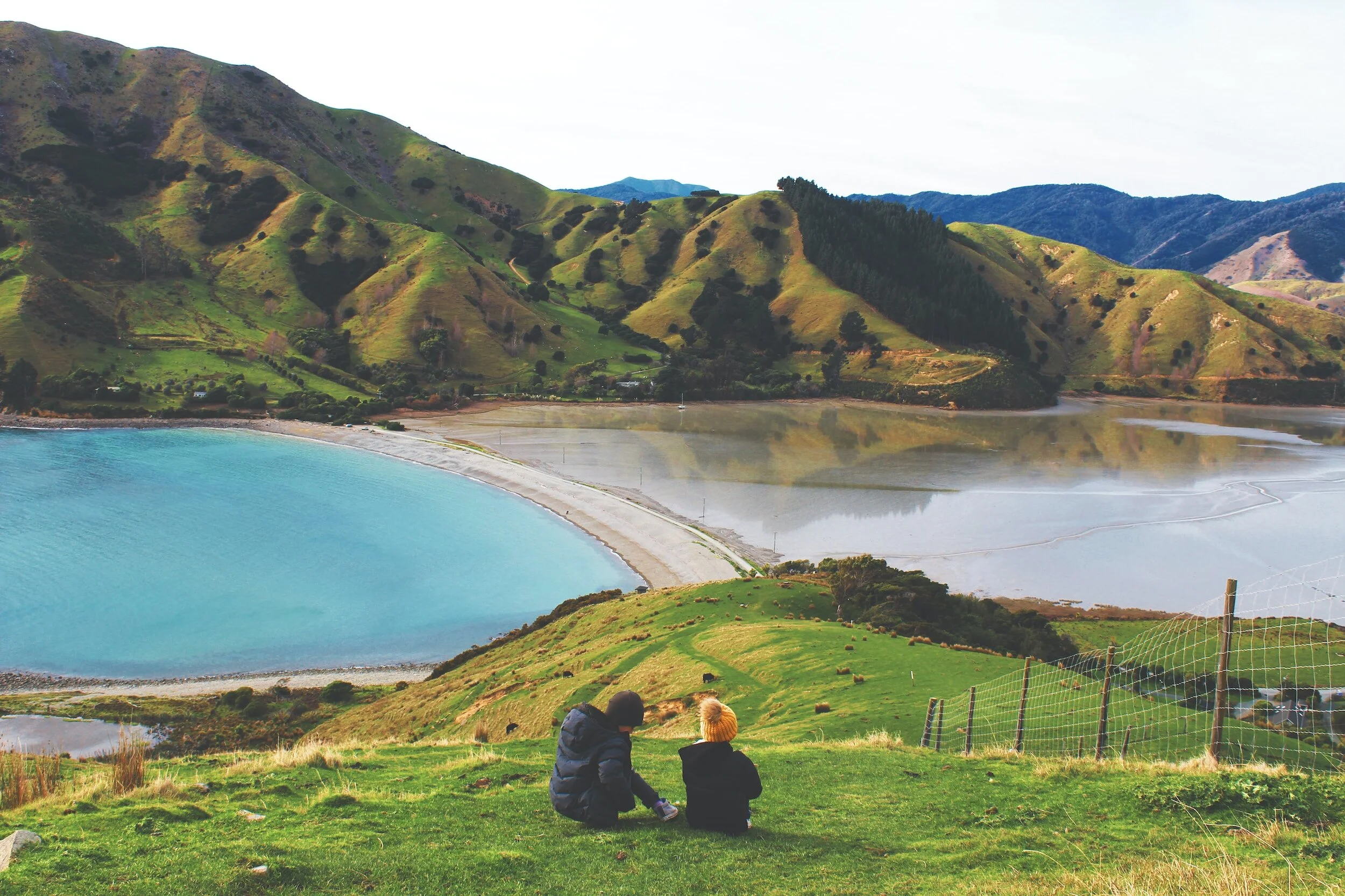 Two children sitting on a grassy hill overlooking a large body of turquoise water with green hills and mountains in the background.