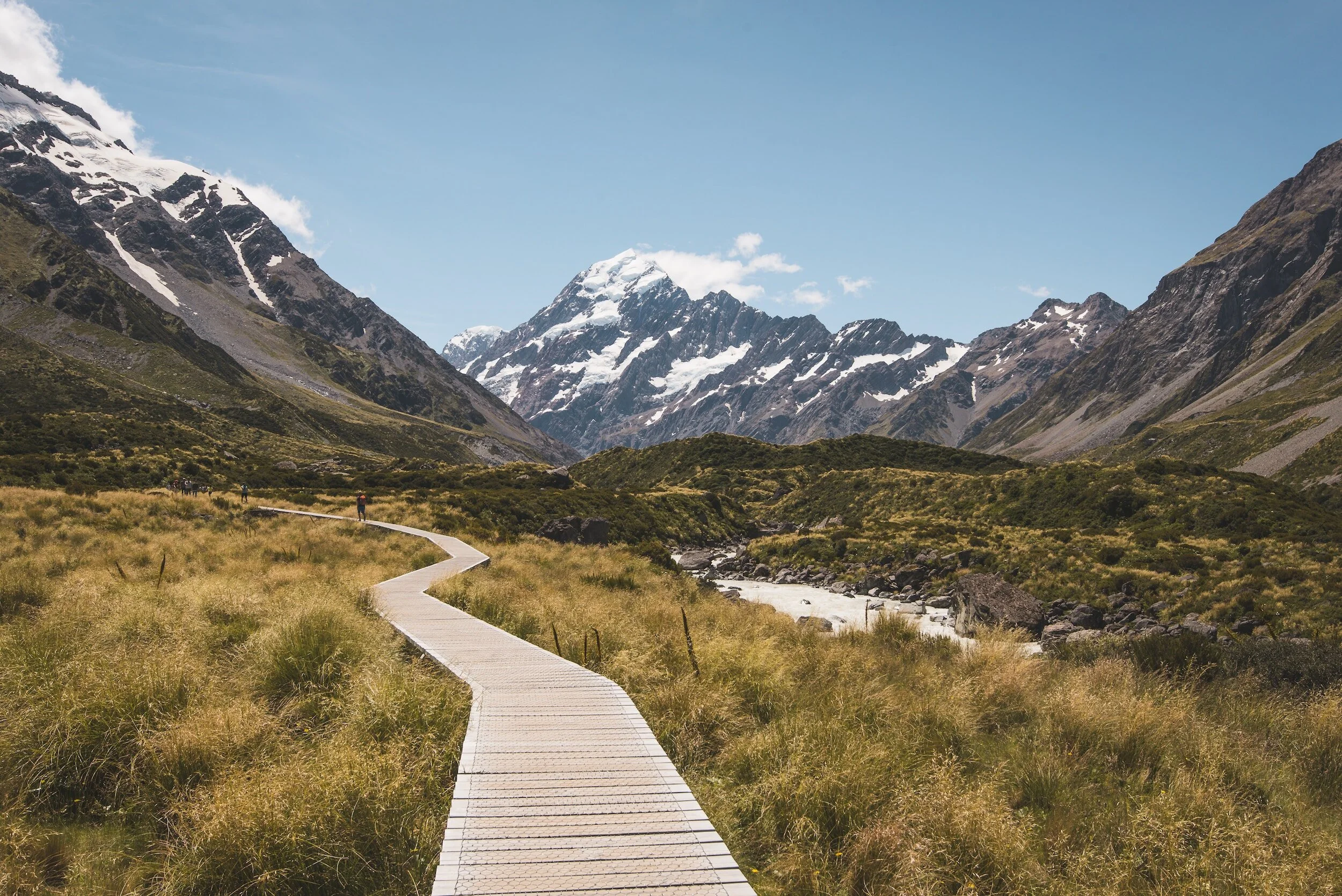 A winding wooden walkway through grassy plains in a mountainous landscape with snow-capped peaks under a blue sky.