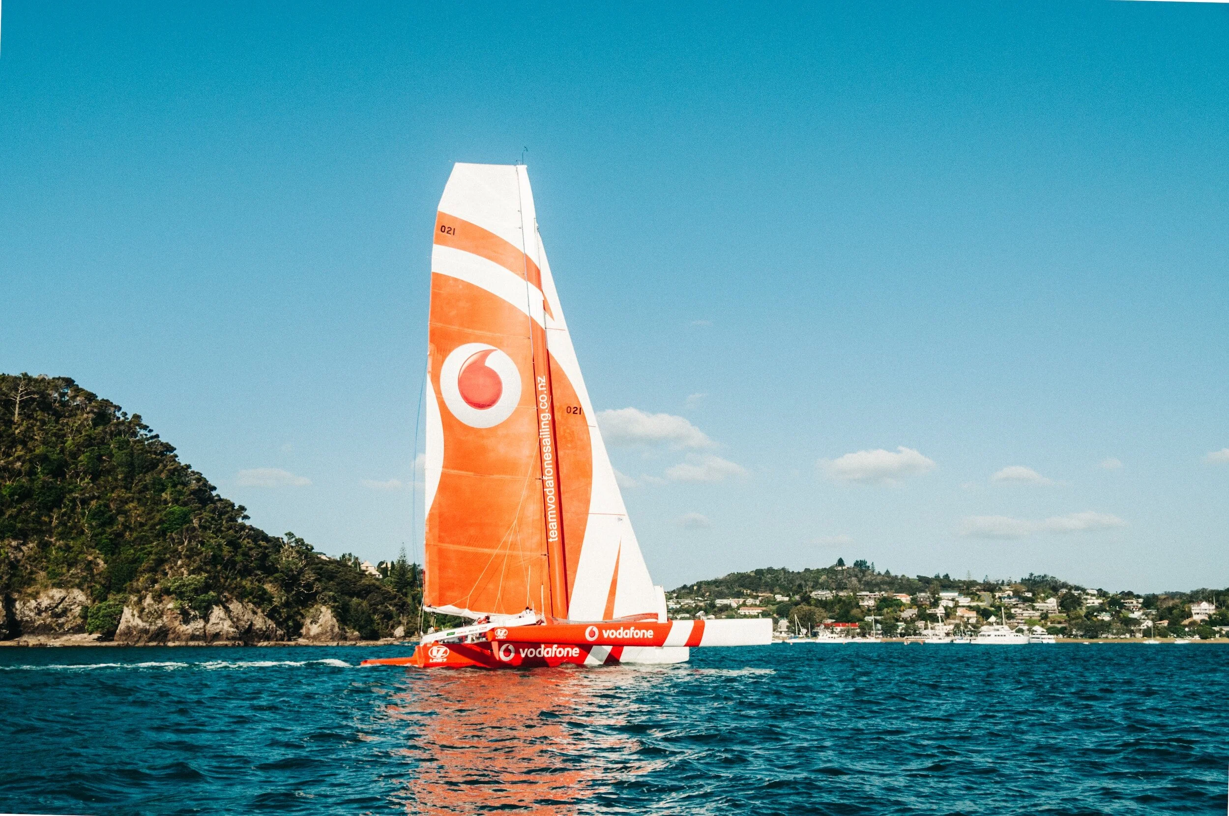 A red and white racing sailboat with Vodafone branding sailing on blue water near a green hillside under a clear blue sky with scattered white clouds.