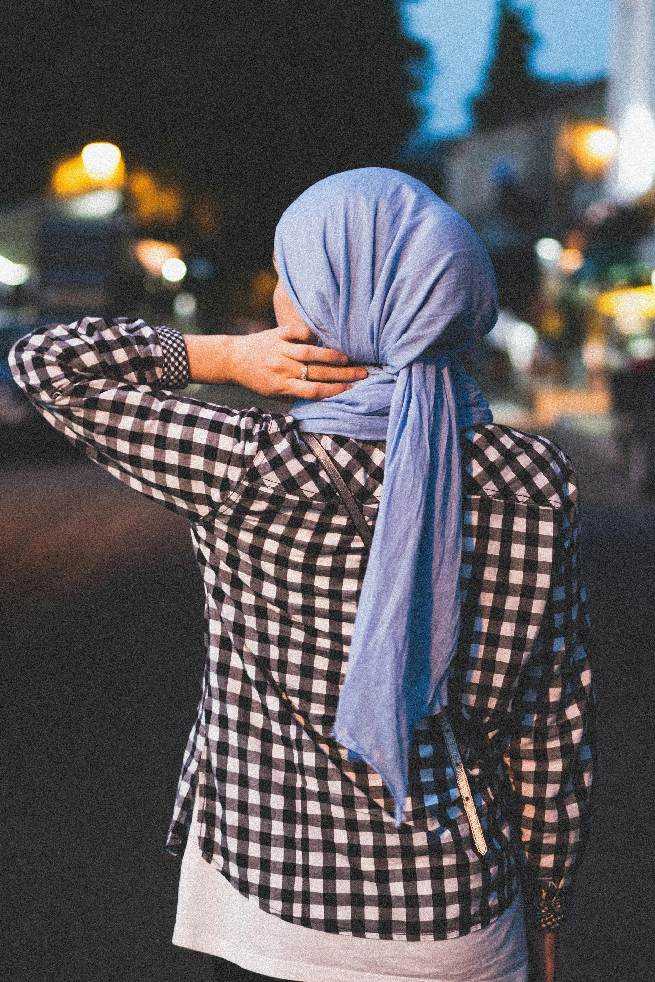 Woman in a checkered shirt and blue headscarf standing on a city street at night, with her hand on the back of her head, surrounded by blurred lights and buildings.