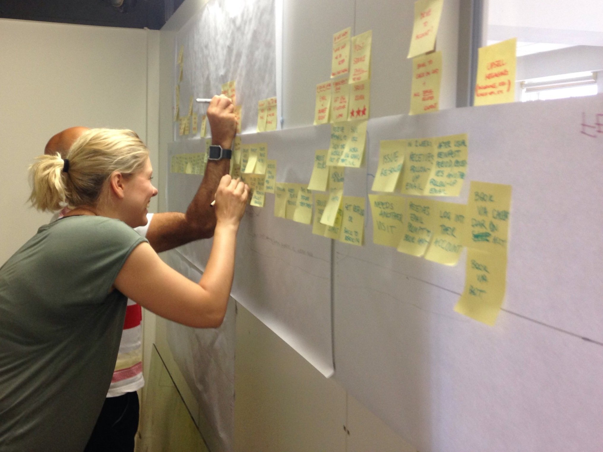 Two people, a woman with blonde hair tied back and a man with bald head, are placing sticky notes on large sheets of paper on a wall, engaged in a brainstorming session.
