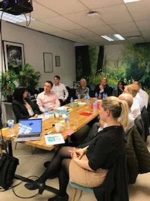 Group of professionals seated around a conference table in a meeting room with laptops, notebooks, and beverages, engaging in a discussion.