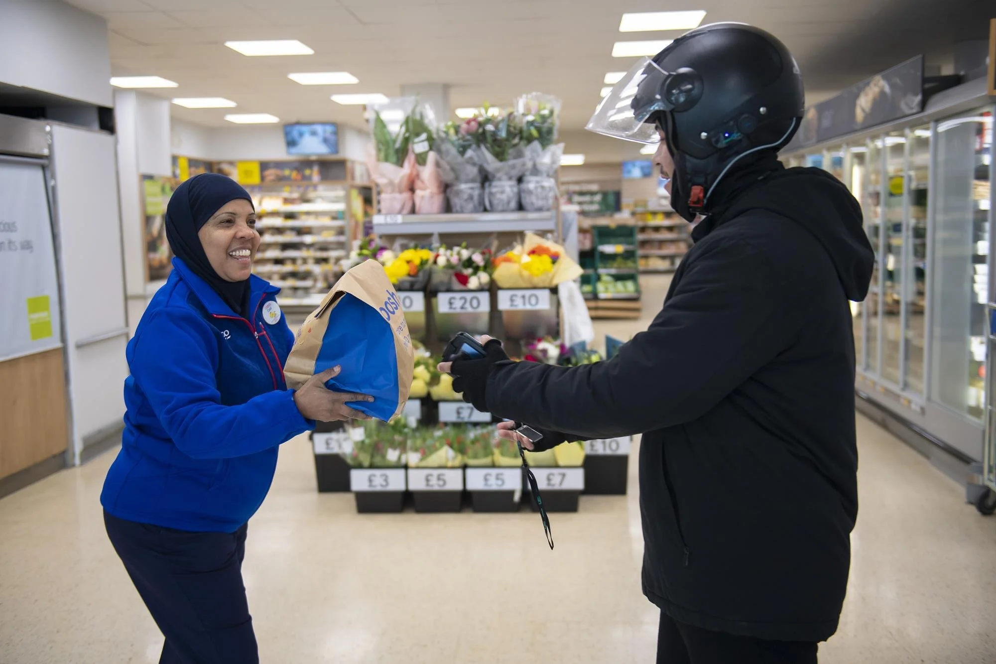 A smiling woman in a blue uniform exchanges a paper bag with a man in a black jacket and helmet at a grocery store checkout. Flower arrangements and shelves with produce are visible in the background.