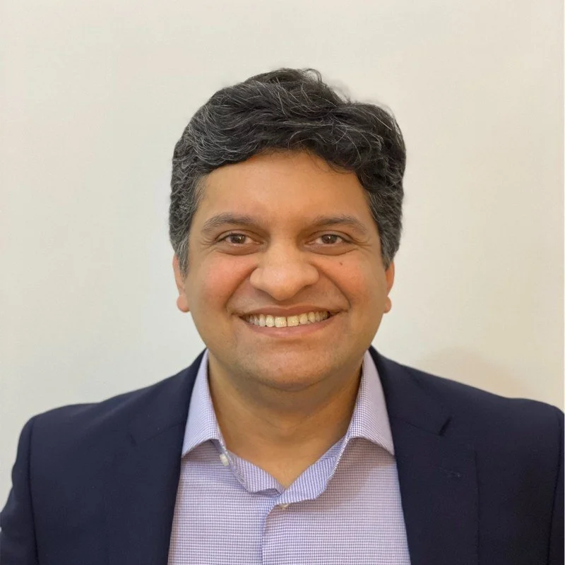 Portrait of a smiling man in a business suit and collared shirt against a plain background.
