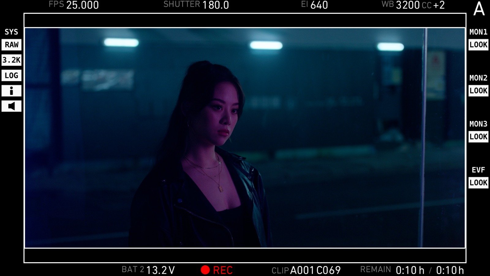A woman with long dark hair, wearing a leather jacket and layered necklaces, standing in front of a glass window at night, illuminated by bluish-purple lighting.