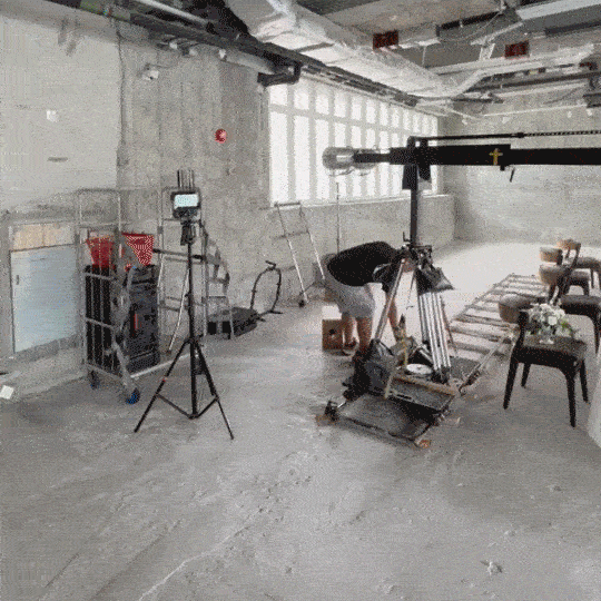 A person operating camera equipment in an unfinished, industrial-style space with concrete floors, exposed ceiling beams, and large windows.