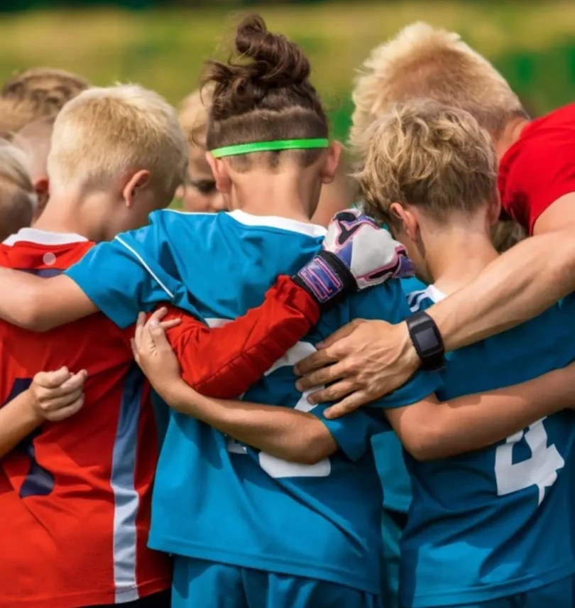 A group of young jakarta international school soccer players in colorful jerseys huddled together in a circle during a game, embracing and supporting each other.