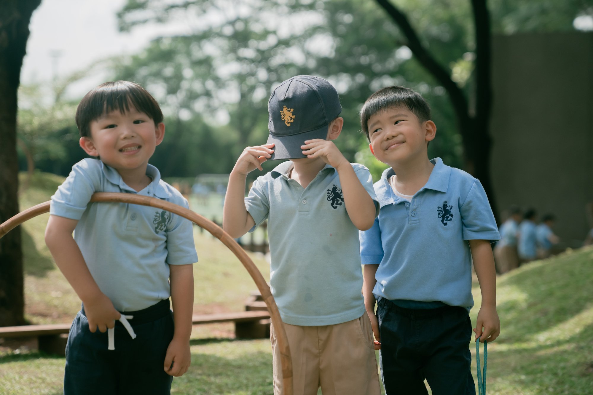 Three young boys at international school jakarta in blue shirts standing outdoors in a park, smiling and looking happy.