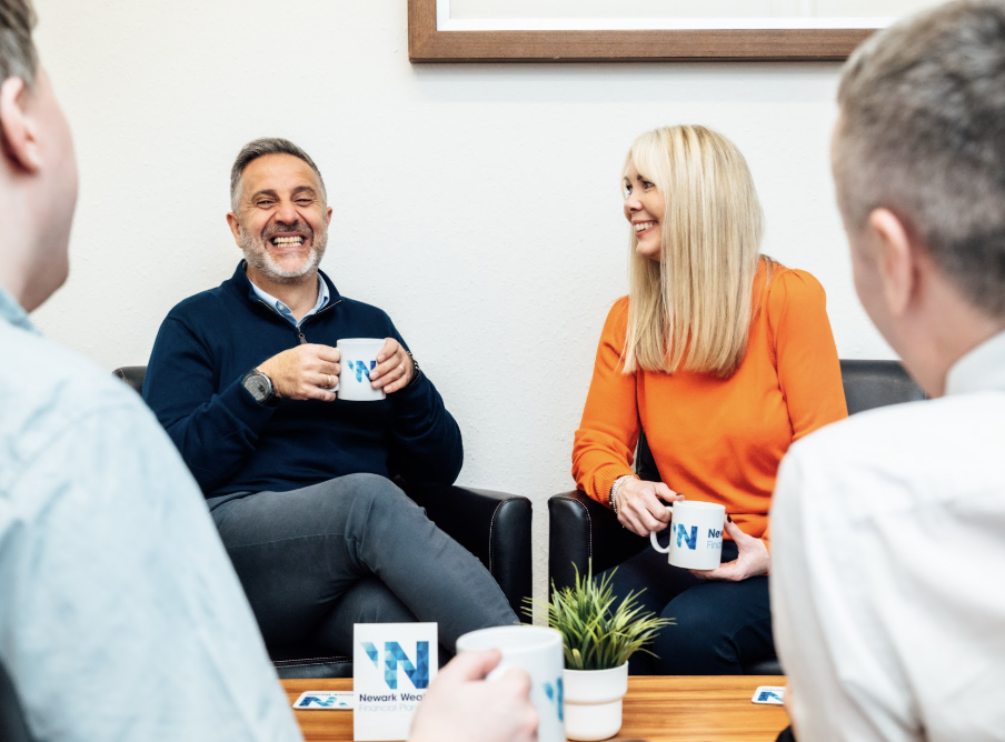 Newark Wealth staff having a meeting in an office, sitting on black chairs, holding coffee mugs, smiling and talking.