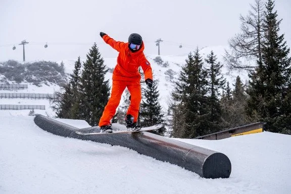 Skiër in oranje kleding glijdt over een bergbekleding in de sneeuw met bomen en skiliften op de achtergrond.