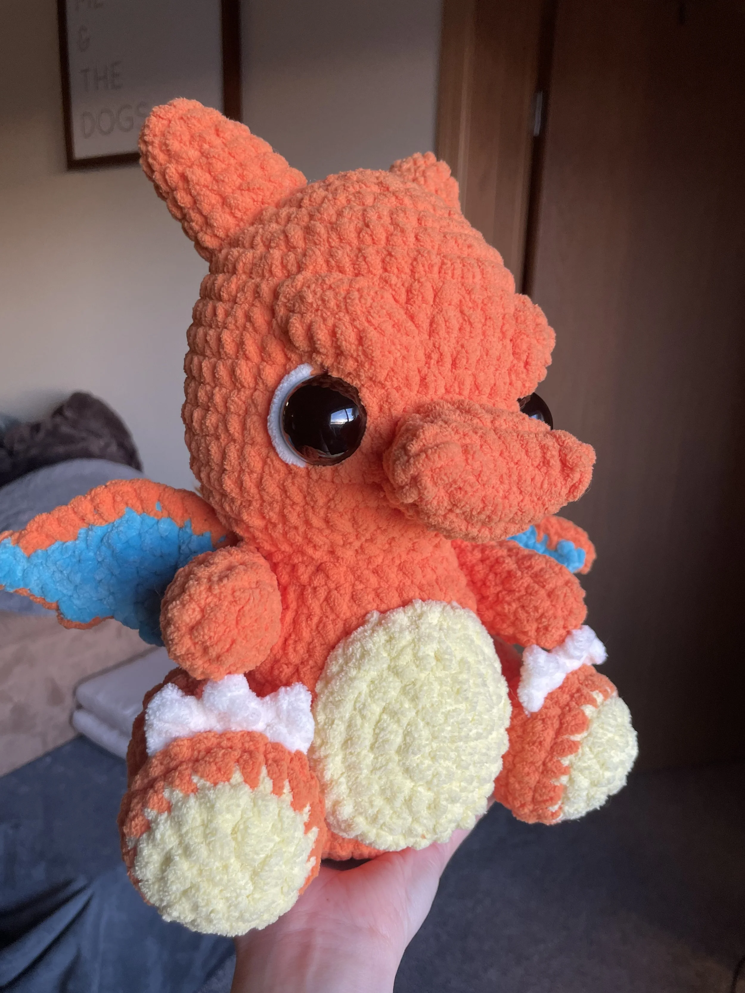 A hand holding a plush toy resembling the Pokémon Charizard, a colorful, cartoon-style dragon with big eyes, orange body, yellow belly, blue wings, and white accents.