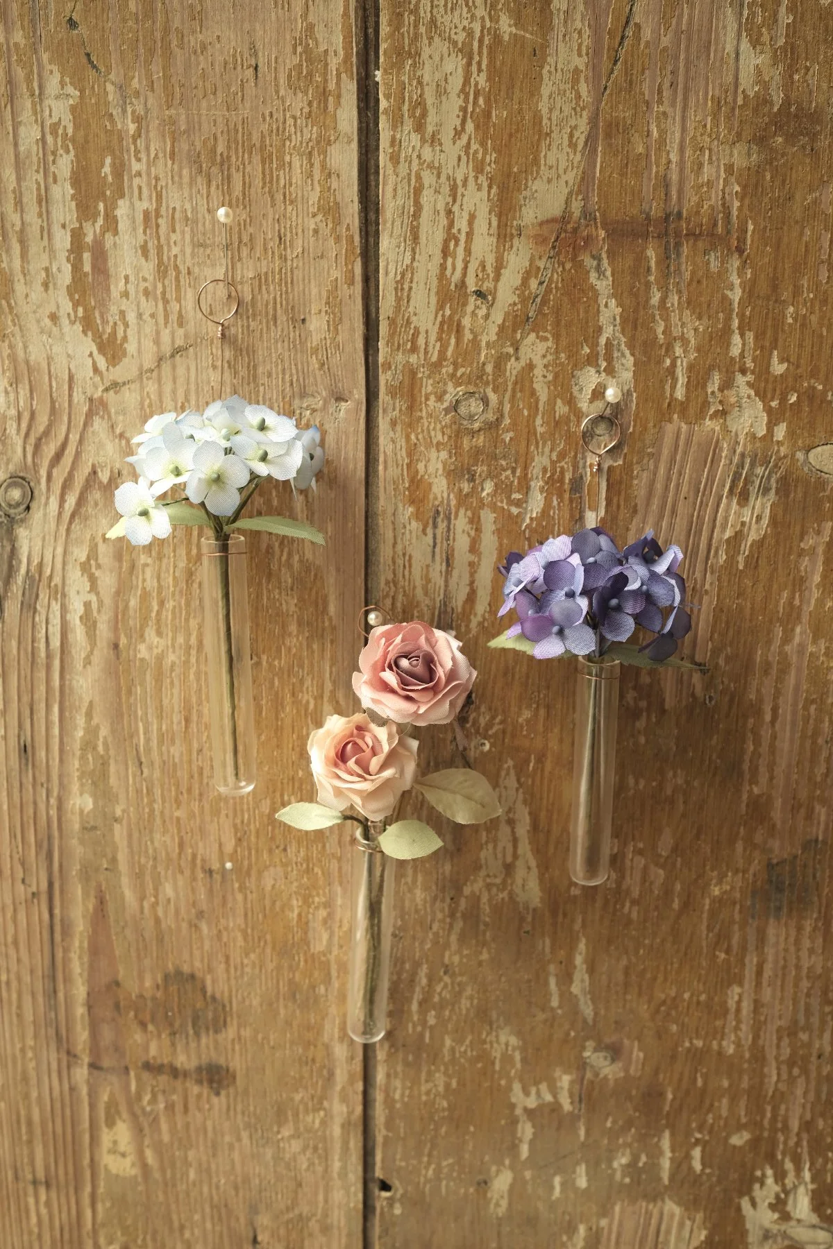 Three artificial flower stems attached to a wooden surface with decorative hooks. The flowers are a white hydrangea, a purple hydrangea, and two pink roses, each with green leaves.