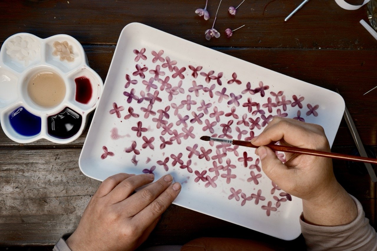 Person painting small pink flowers on a white tray with watercolor paints, including various colors in a palette, on a wooden surface.