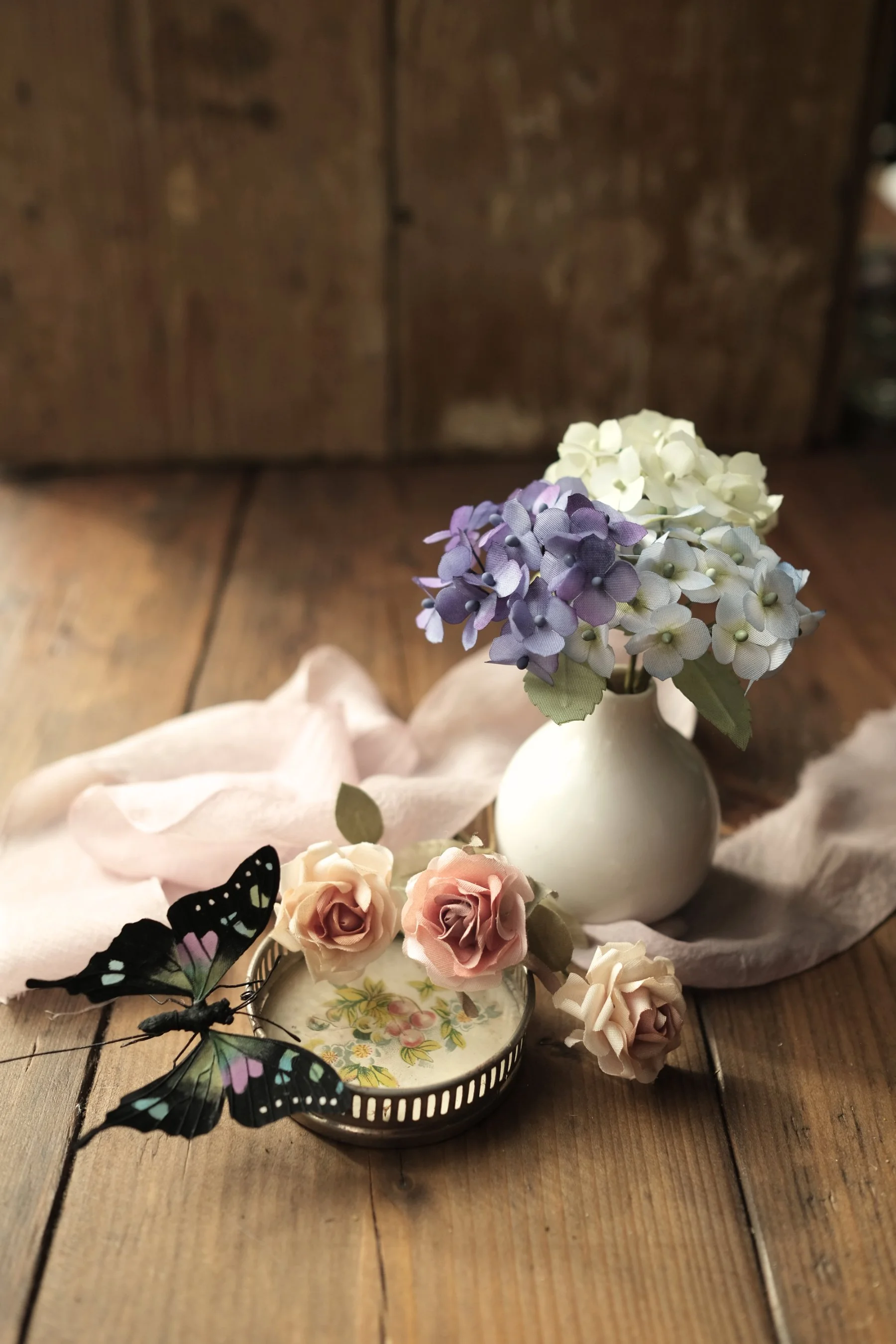 A white vase with purple and white hydrangea flowers, placed on a wooden surface with a pink cloth. There are pink and white roses and a butterfly decoration nearby. A decorative tray with floral patterns is underneath some flowers. The background is a dark wooden wall.