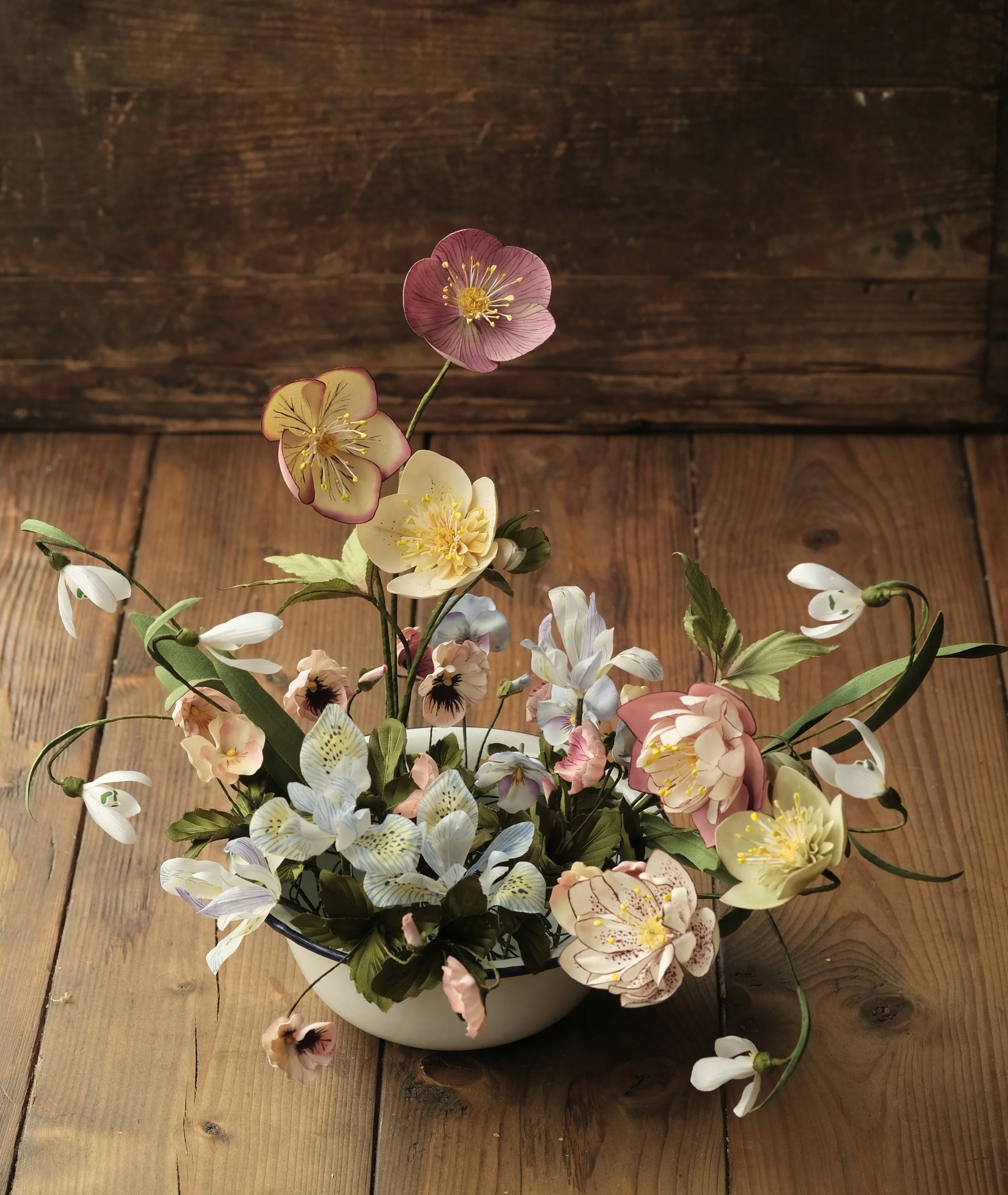 A ceramic vase filled with artificial flowers, including pink, white, and purple blossoms, placed on a wooden surface with a wood-paneled background.