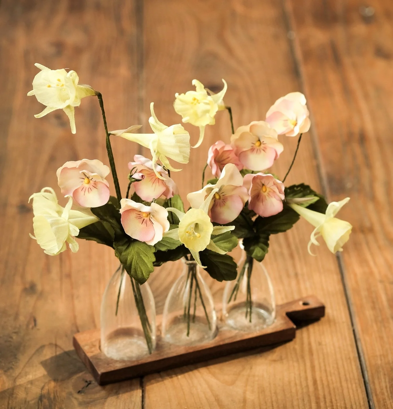 A set of three small glass vases with yellow and pink artificial flowers, placed on a wooden table.
