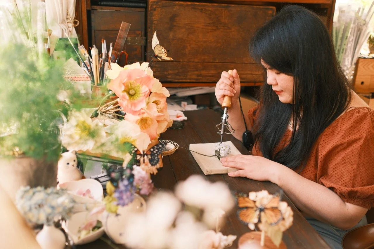 Woman crafting jewelry at a wooden workspace decorated with flowers and butterflies, surrounded by art supplies and decorative items.