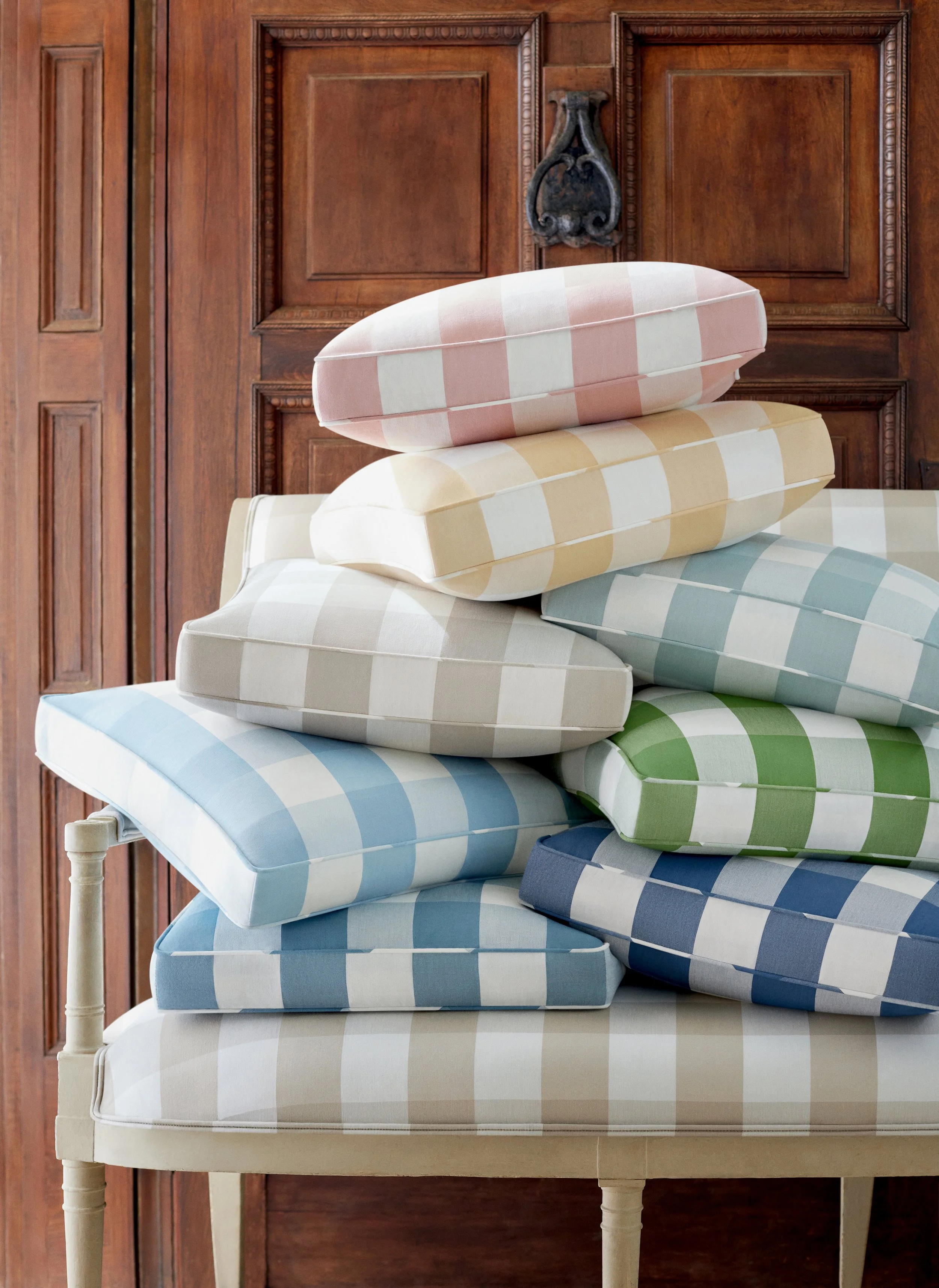 Stacked pastel-colored checked cushions in pink, yellow, blue, and green, on a wooden chair in front of wooden paneling.