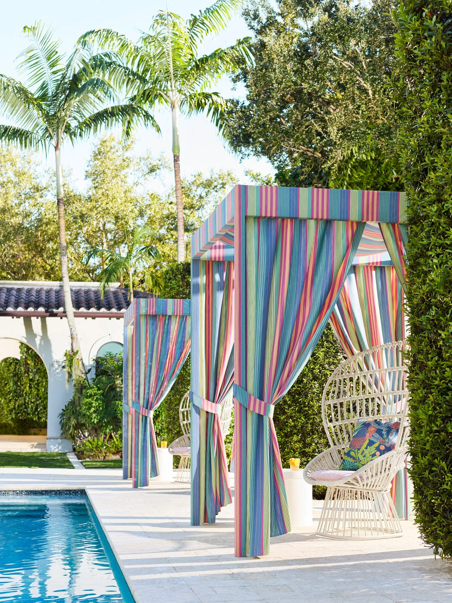 Colorful striped canopy near a swimming pool with white wicker chairs and lush greenery.