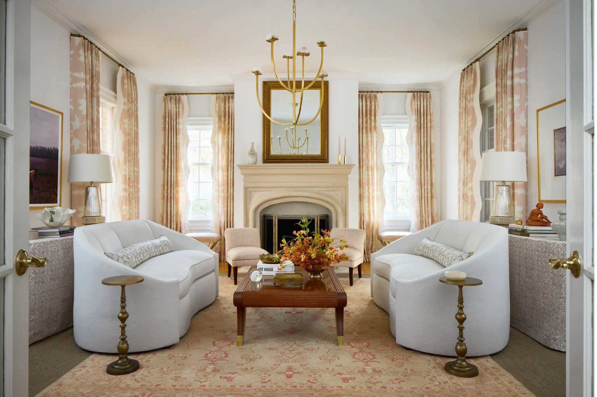 A symmetrical living room with beige and white furniture, a fireplace in the center, floral curtains, and a chandelier hanging from the ceiling.