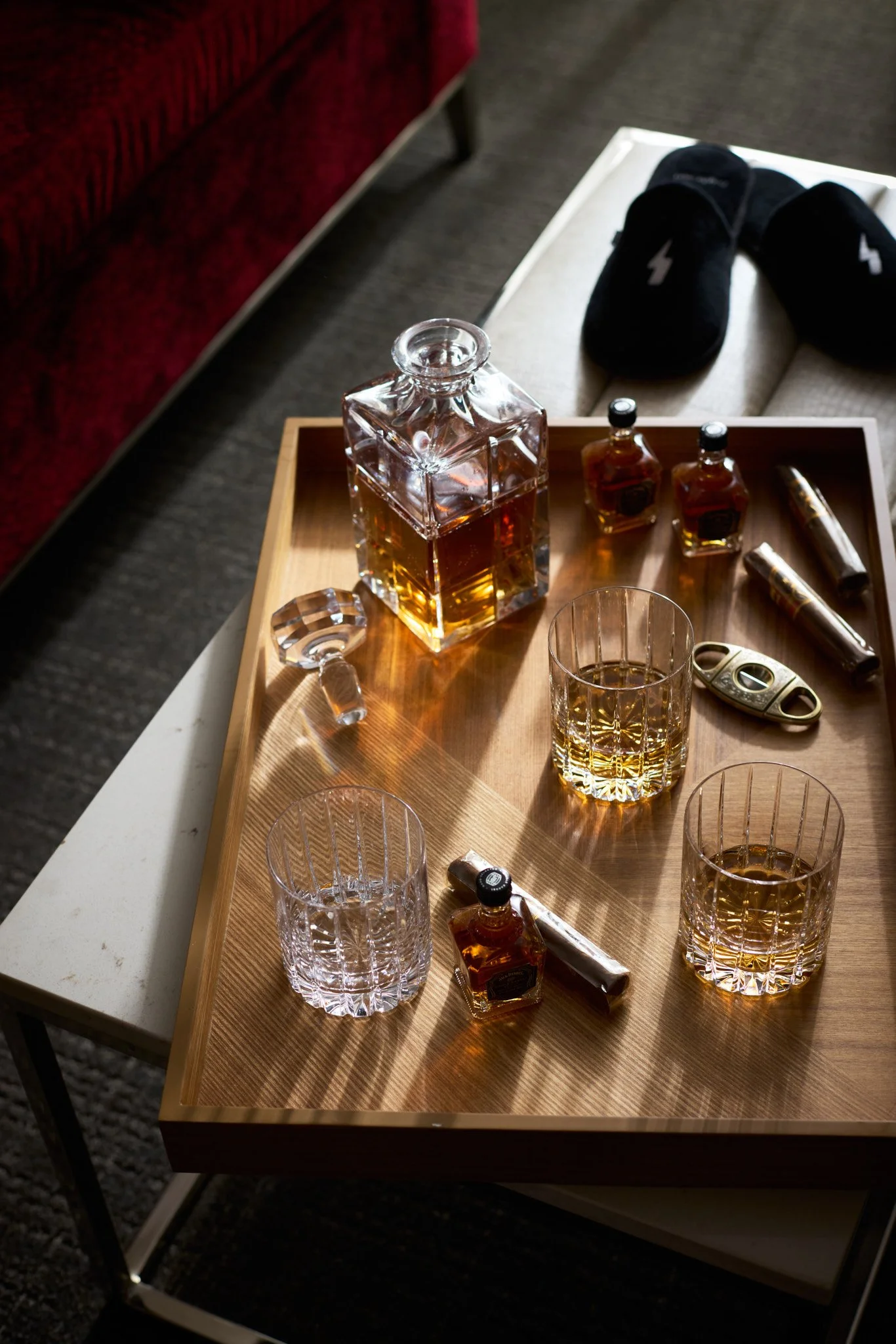 A wooden side table with a large glass decanter of whiskey, two small whiskey bottles, and two empty glasses, with a pair of black slippers and a shoehorn on a beige ottoman in the background.