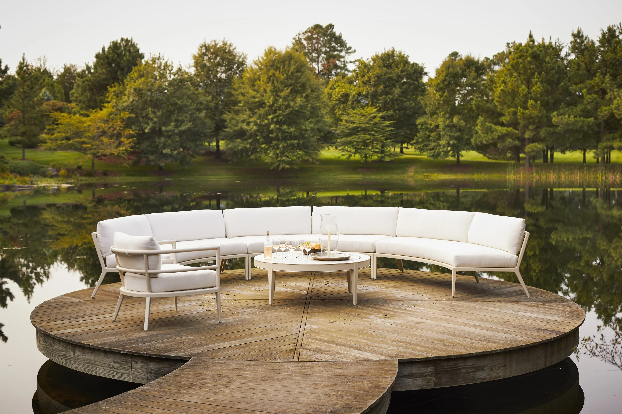A white outdoor sectional sofa with a small round table holding drinks, situated on a wooden deck over a pond with a lush green park and trees in the background.