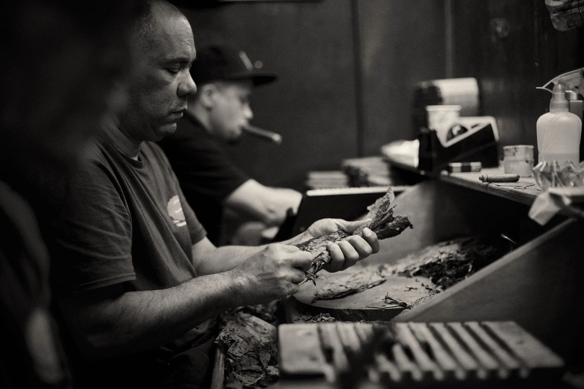 Two men sitting at a bar, one of them peeling or preparing a fish, with various items like bottles and paper on the counter.